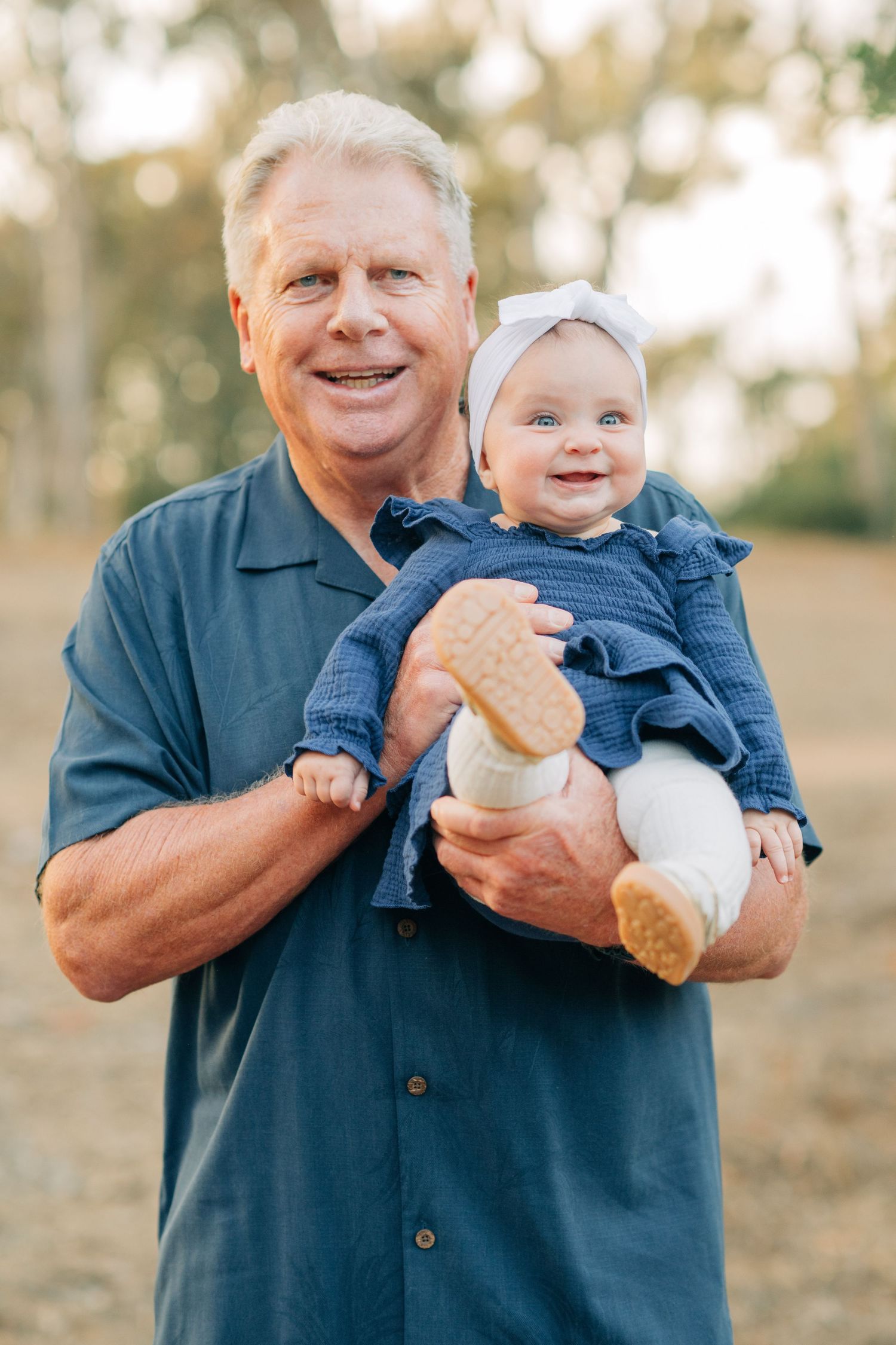 Smiling older man holding a happy baby girl in a blue outfit with a white bow outdoors.