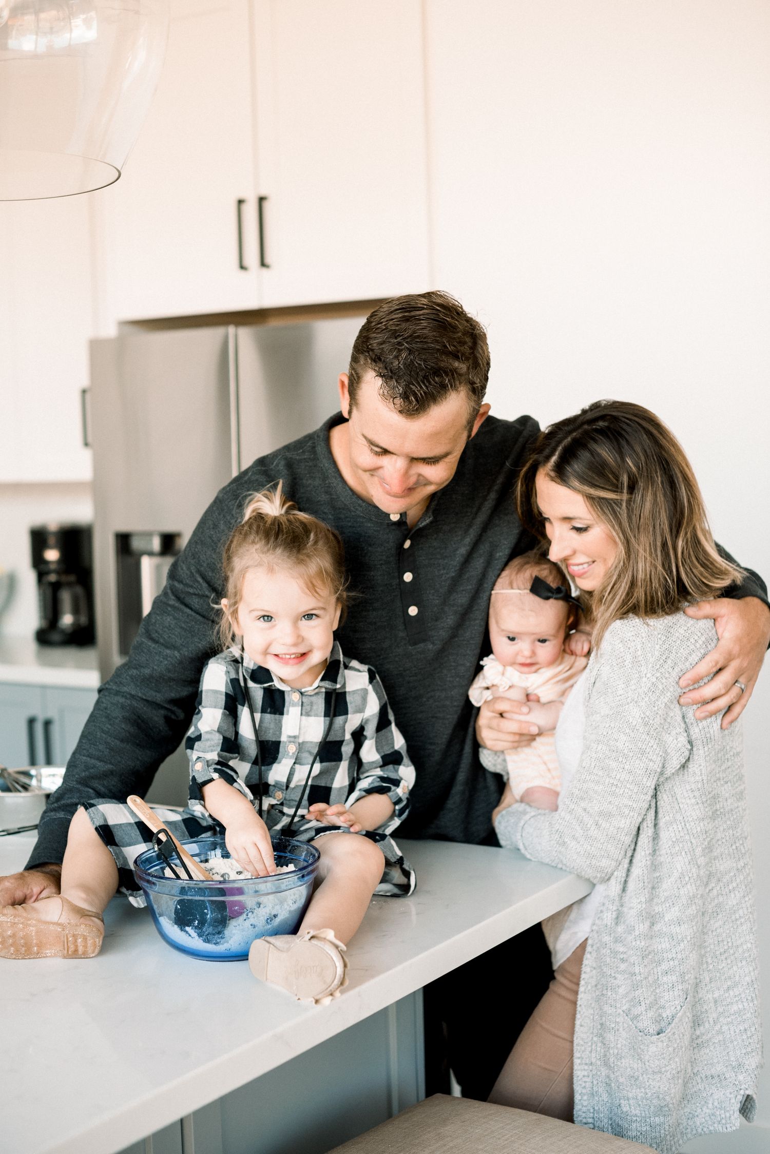 Happy family of four gathered around kitchen counter, parents smiling at toddler mixing bowl while mom holds newborn baby.