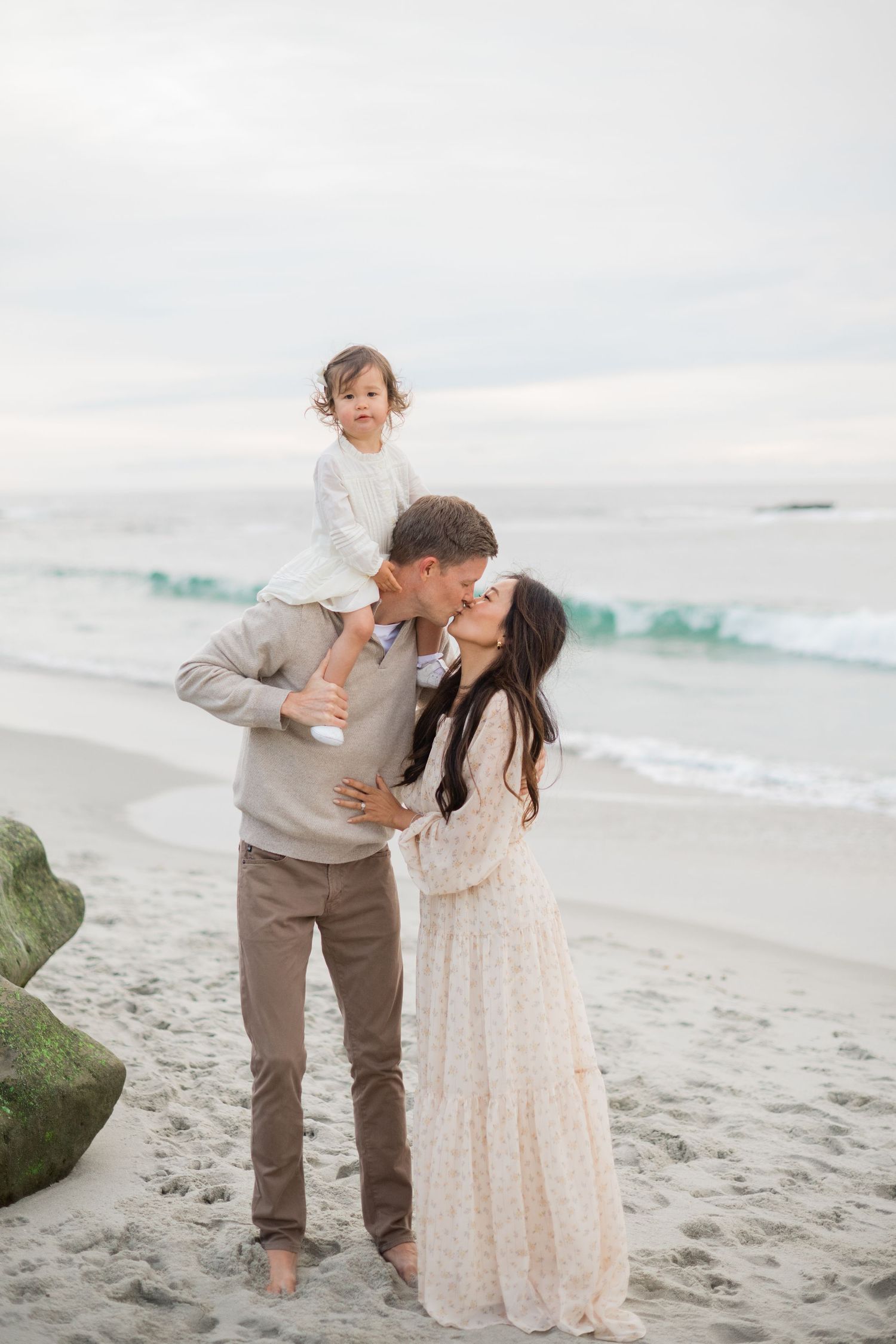 A young family shares a kiss on a sandy beach, with a toddler held up by the father near ocean waves.