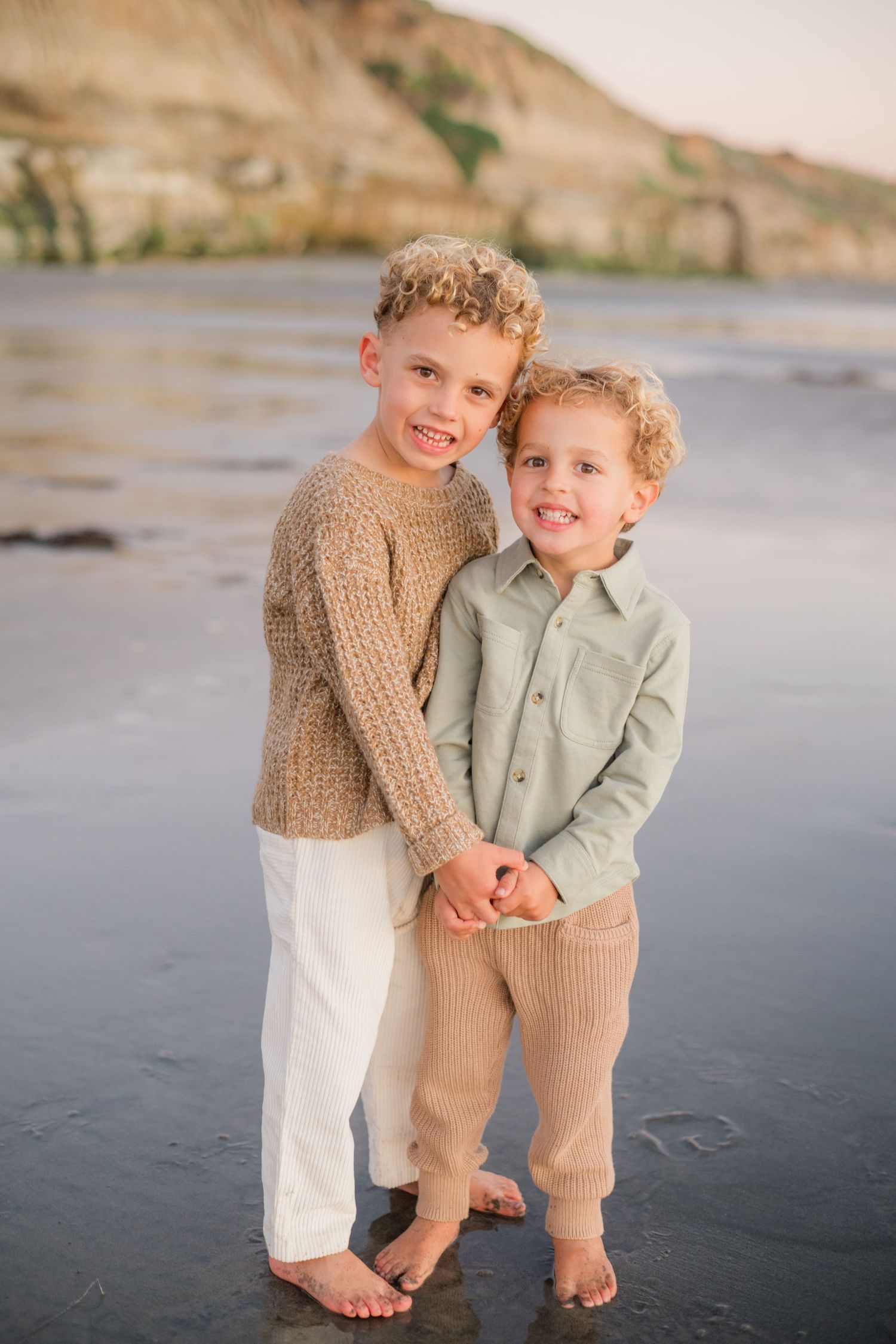 Two young brothers holding hands and smiling on a sandy beach with cliffs in the background.