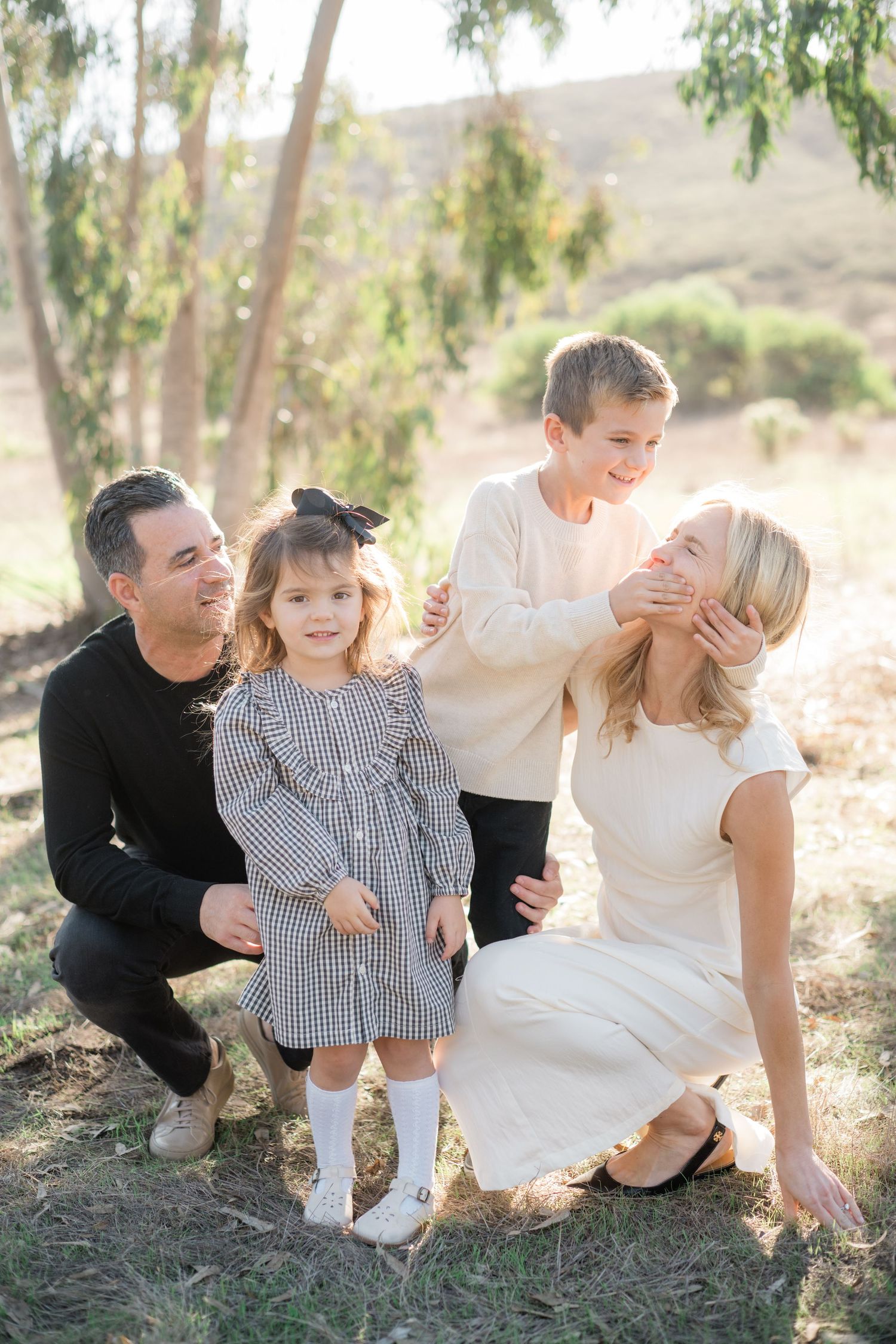 Happy family of four posing outdoors, parents with young daughter in plaid dress and son among trees.