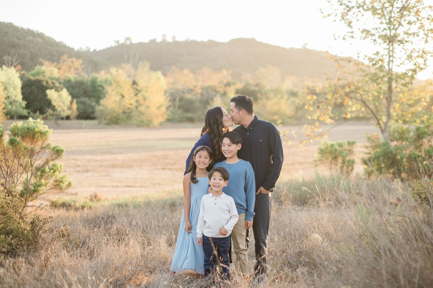 Family of five posing in a golden field with autumn trees and hills in the background during sunset.