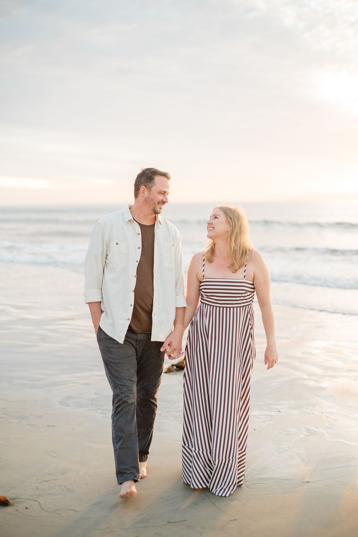 A smiling couple holds hands while walking barefoot along a sandy beach at sunset near the ocean.