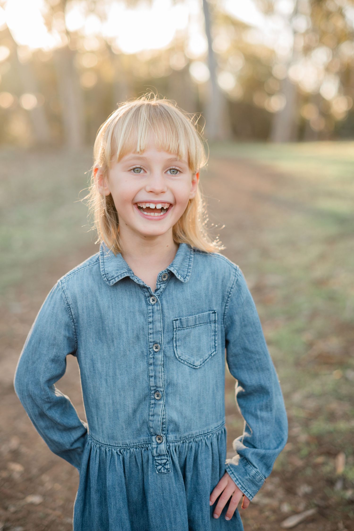 Happy young blonde girl in denim dress laughing with hands on hips in a sunny outdoor park.