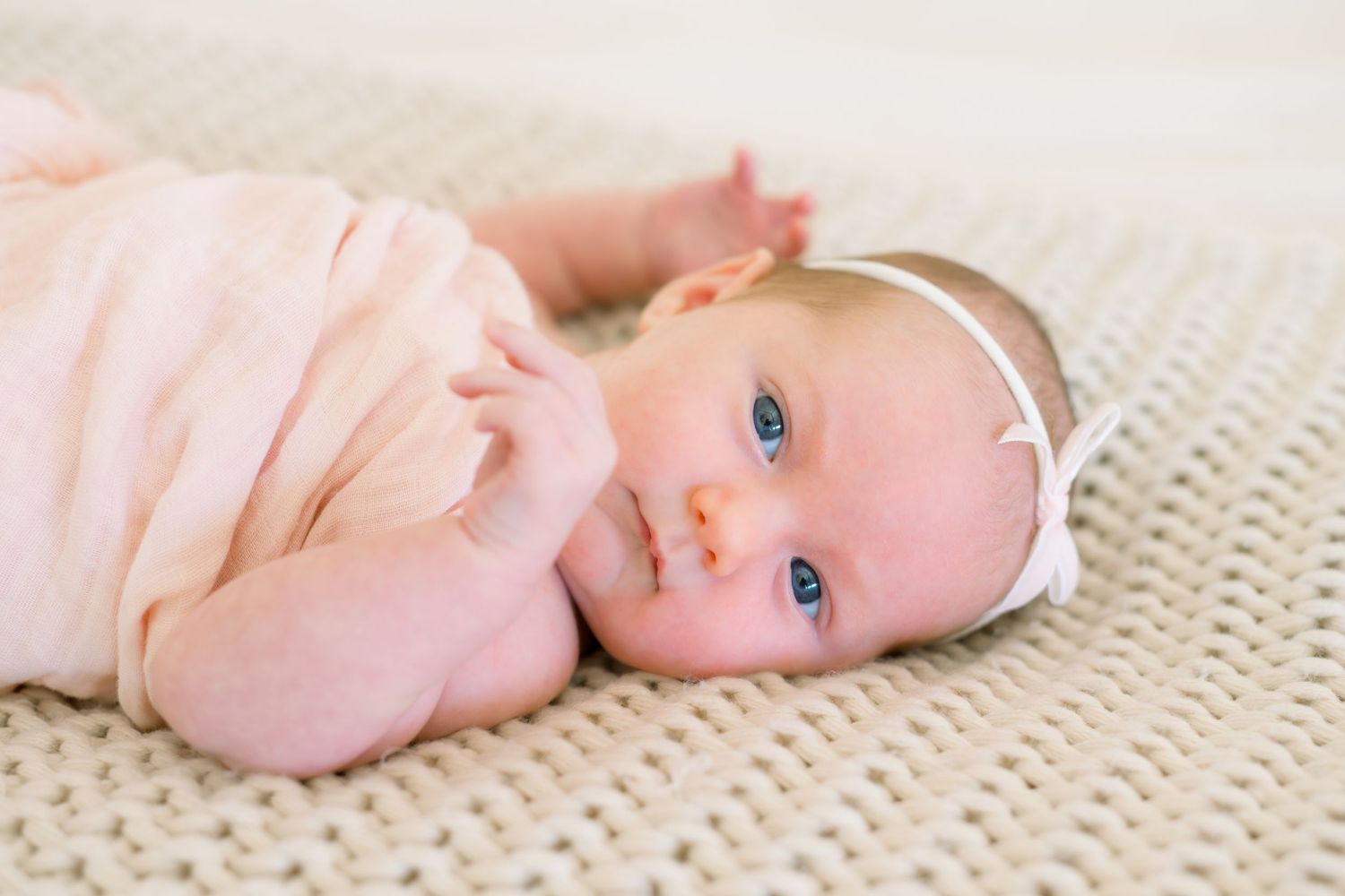 A cute baby girl with blue eyes wearing a pink outfit and white headband lying on a knit blanket.