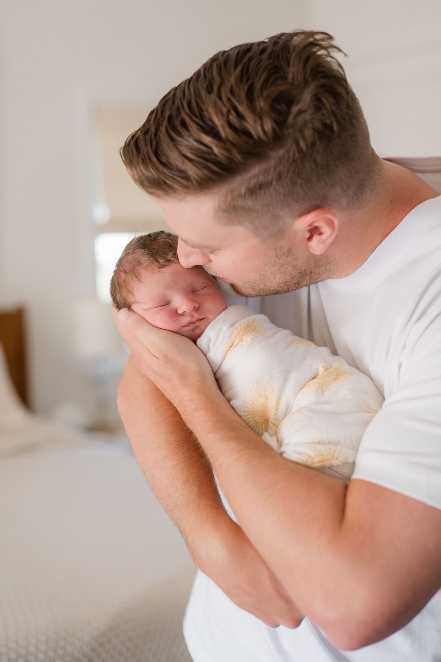 Father tenderly kissing sleeping newborn baby wrapped in white swaddle blanket indoors.