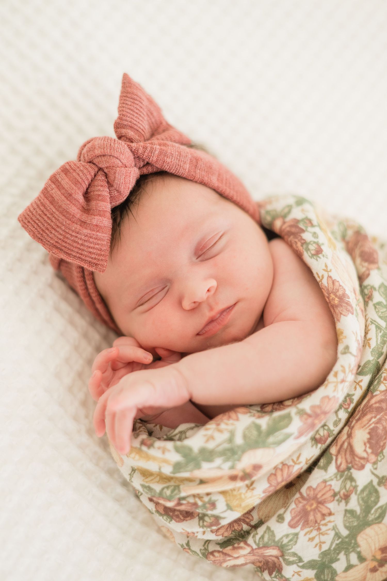 Sleeping newborn baby wrapped in floral swaddle with pink bow headband on white background.