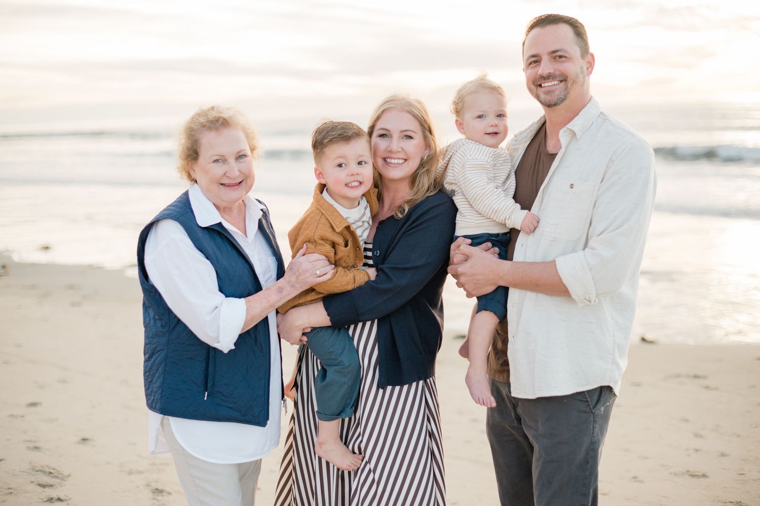 Family of five smiling on a sandy beach, including grandmother, parents, and two young children, with ocean waves in the background.