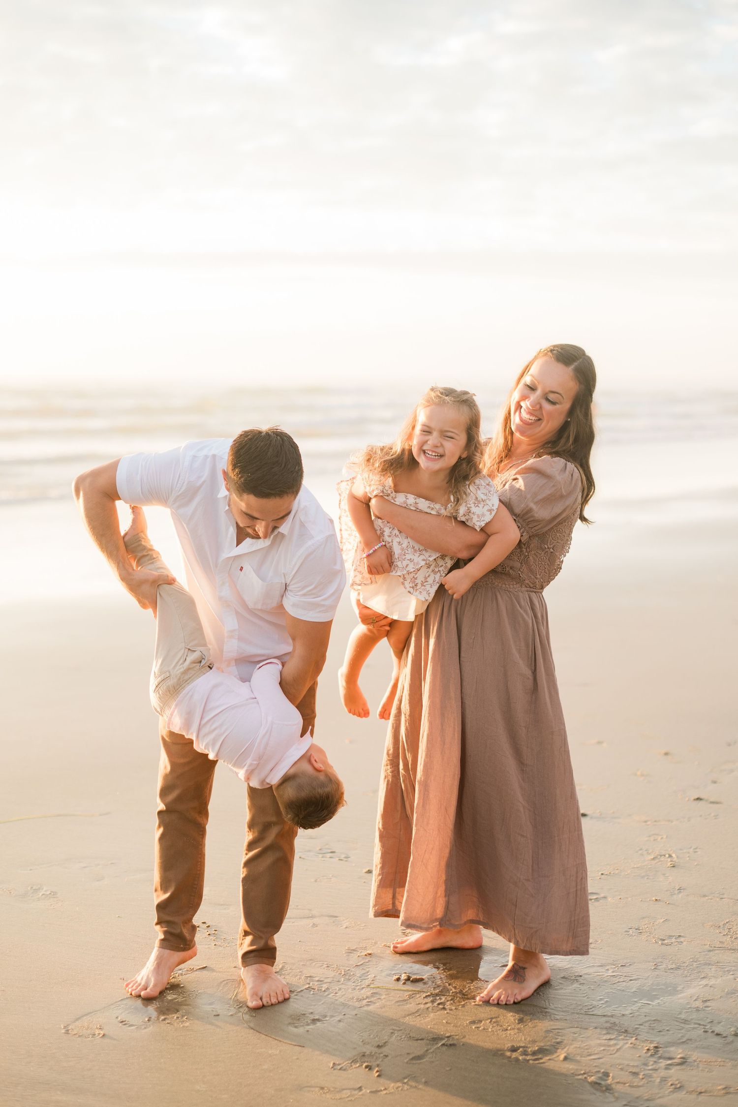 Family of four laughing and playing on a sandy beach at golden hour, parents swinging toddler while daughter smiles.
