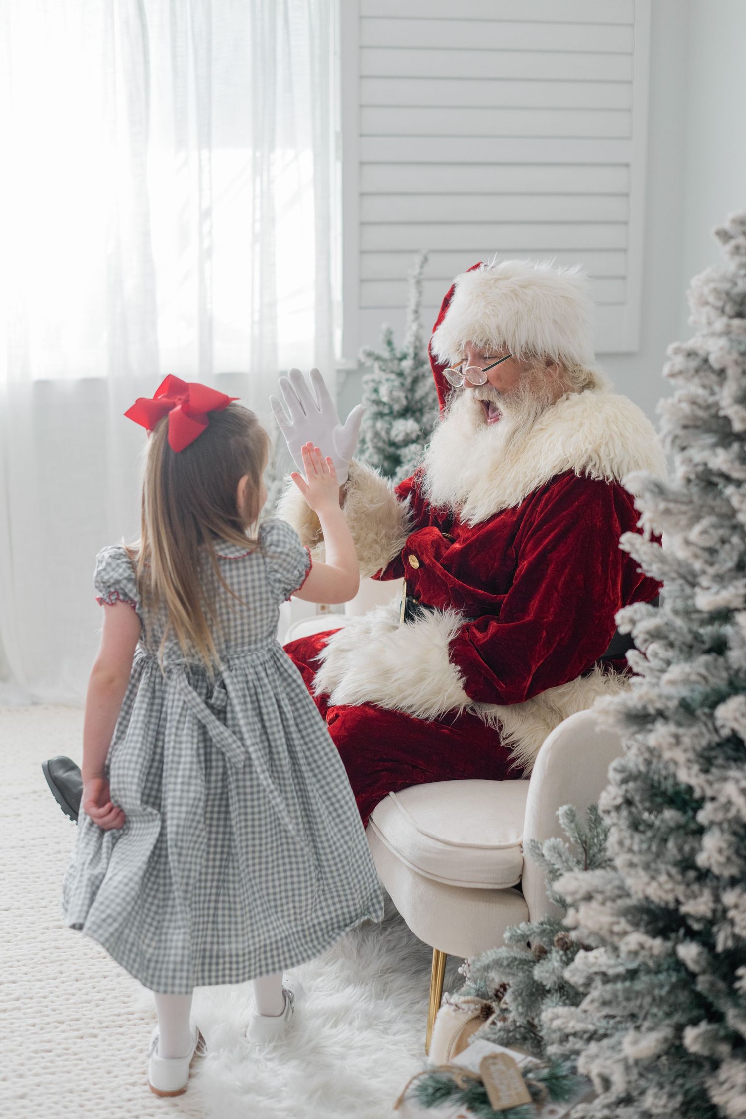 A little girl in a plaid dress gives Santa Claus a high five beside a snow-flocked Christmas tree.