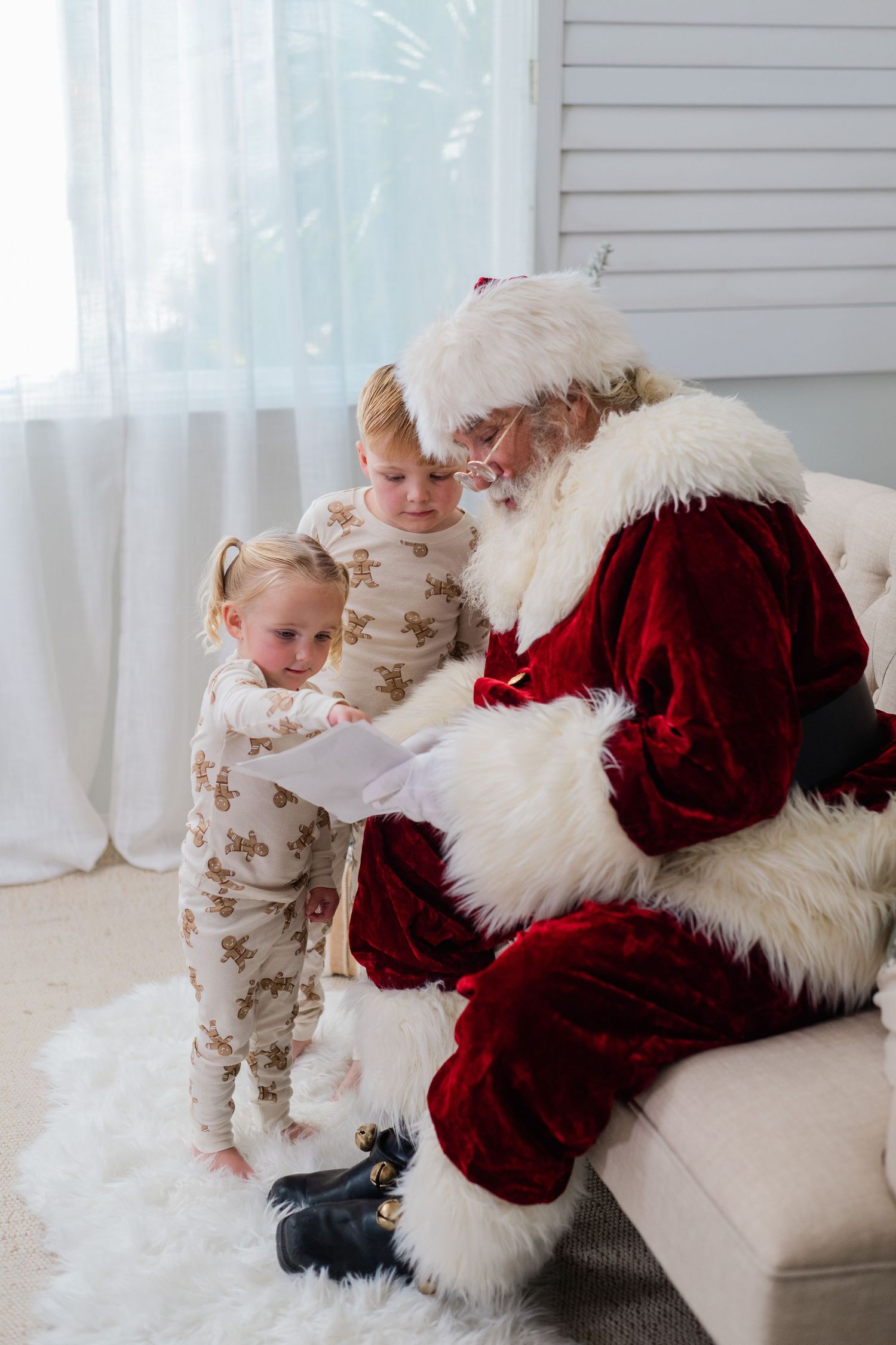 Two young children in holiday pajamas sitting with Santa Claus on a couch, reading a Christmas wish list.