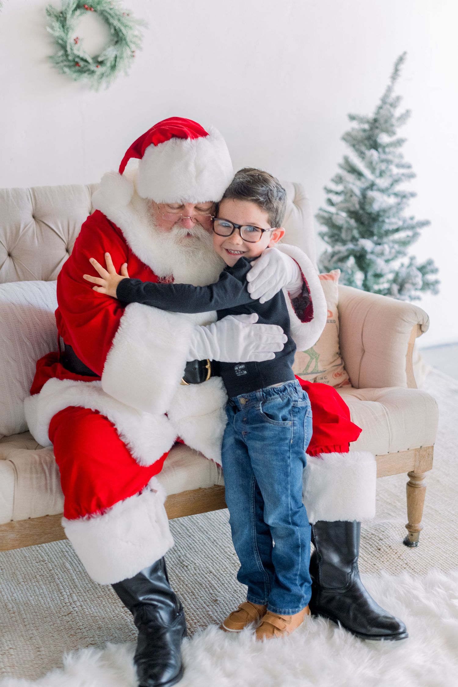 A young boy wearing glasses hugs Santa Claus warmly while sitting on a couch with a Christmas tree nearby.