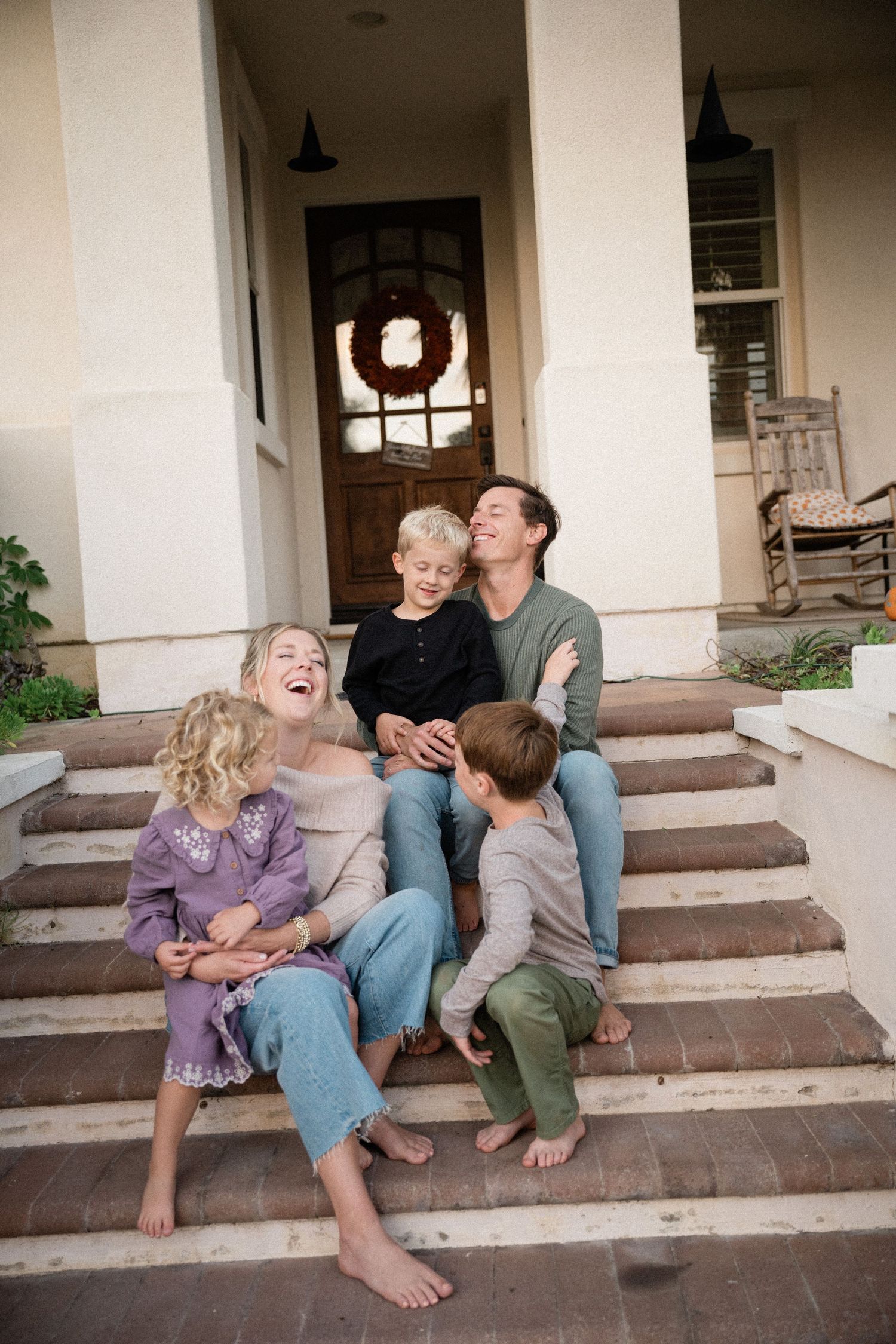 Happy family laughing together on front porch steps of a home, parents with two young children enjoying a candid moment.