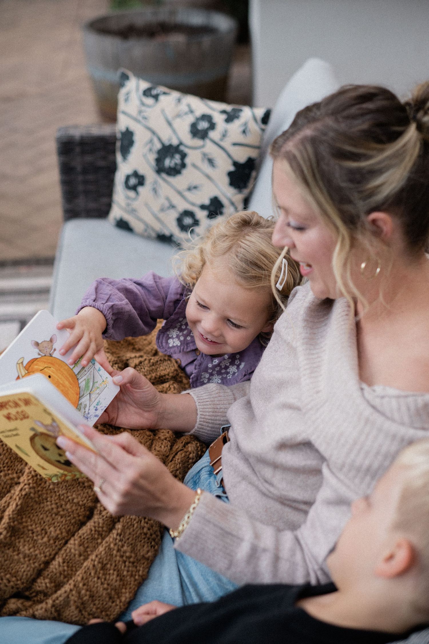 Blonde mother in cream sweater reading a colorful picture book with her two young children outdoors.