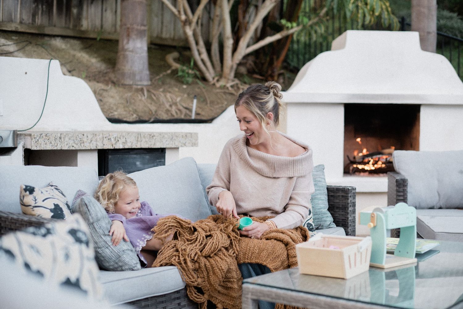 Mother and toddler relaxing on outdoor sofa near fireplace with brown blanket and toys.
