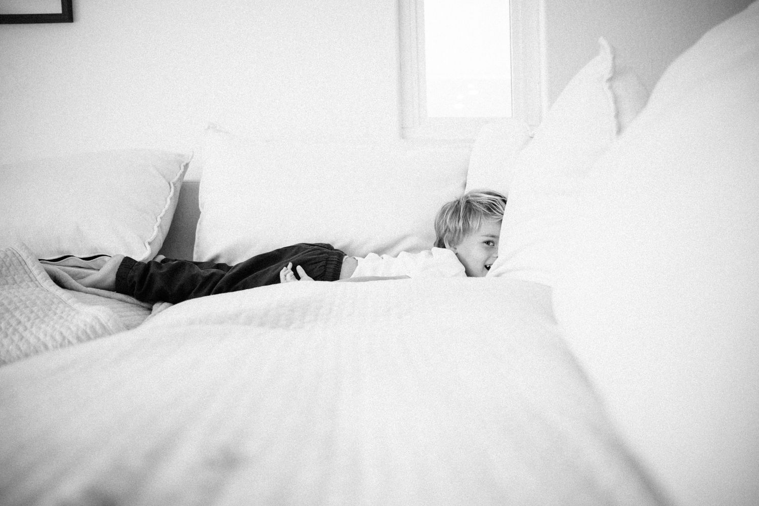 Black and white photo of a young child lying on a white bed, peeking out from behind fluffy pillows.