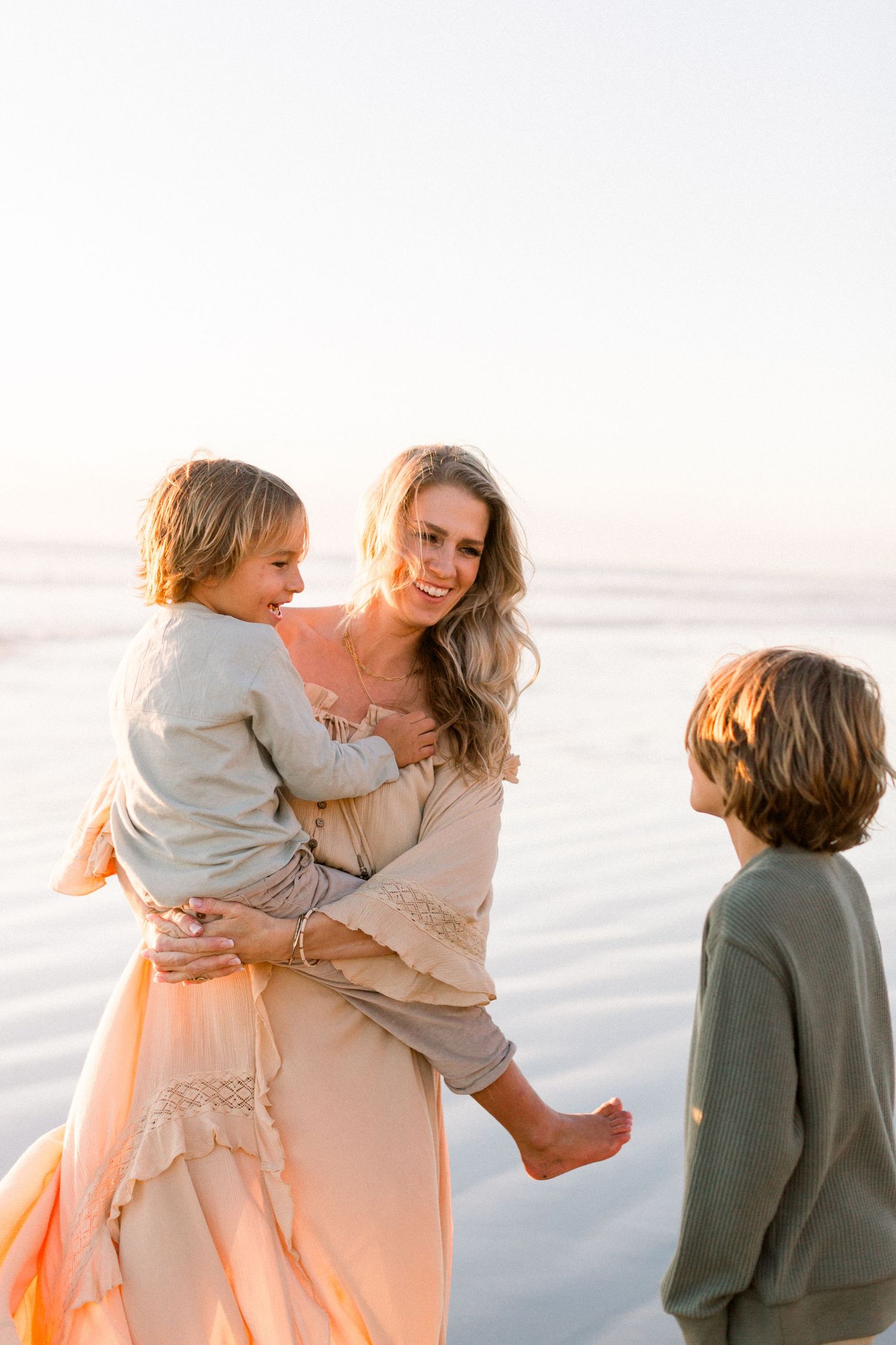 A smiling woman in a peach dress holds a child while another child stands nearby on a sunlit beach.