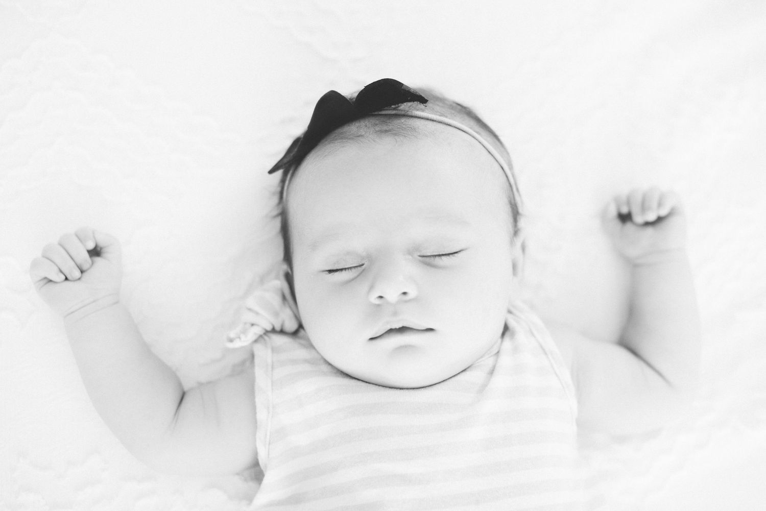 Peaceful newborn baby girl wearing a black bow headband sleeping on a white blanket.
