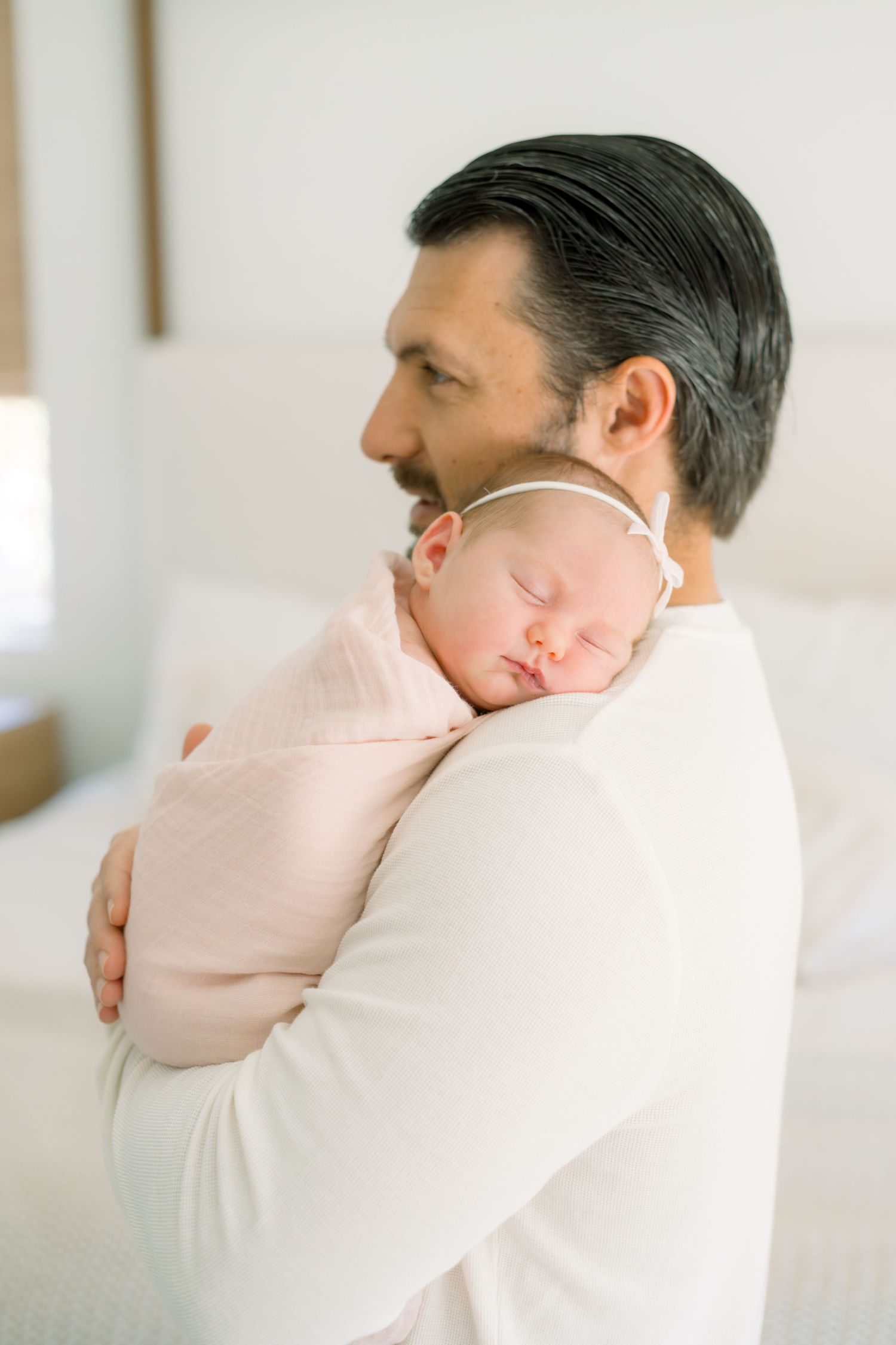 Father in white shirt tenderly holds sleeping newborn baby wrapped in pink swaddle blanket.