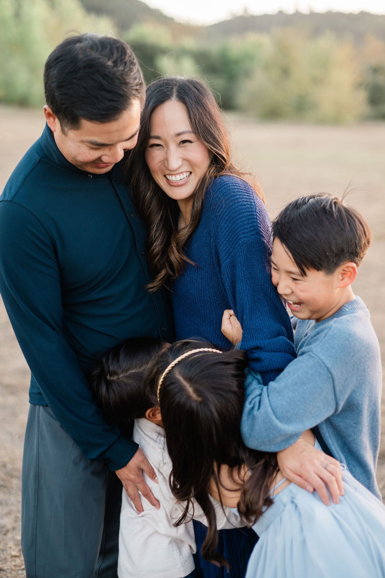 A joyful Asian family of four shares a warm group hug in an outdoor field during golden hour.