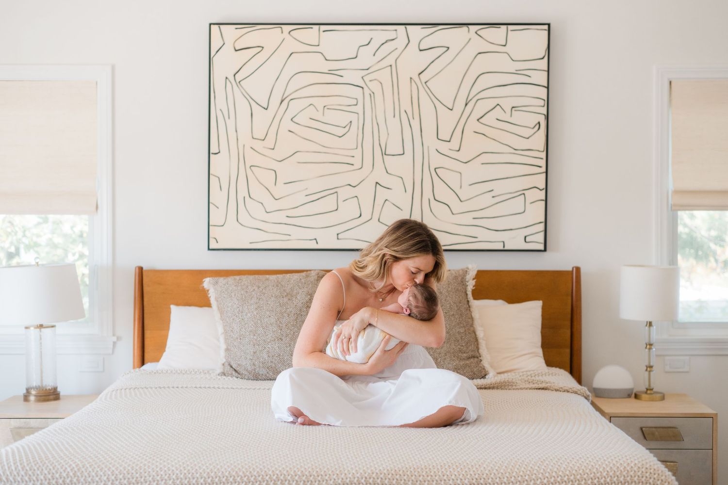 A mother sits on a white bed, tenderly cradling her newborn baby, with abstract wall art hanging above.