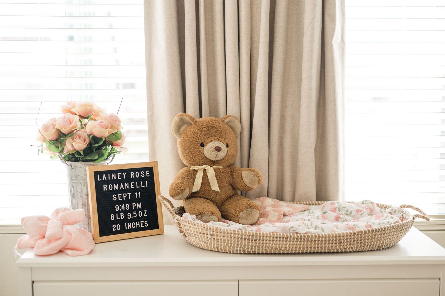 Newborn baby announcement letter board with teddy bear and floral bassinet on white dresser in nursery.