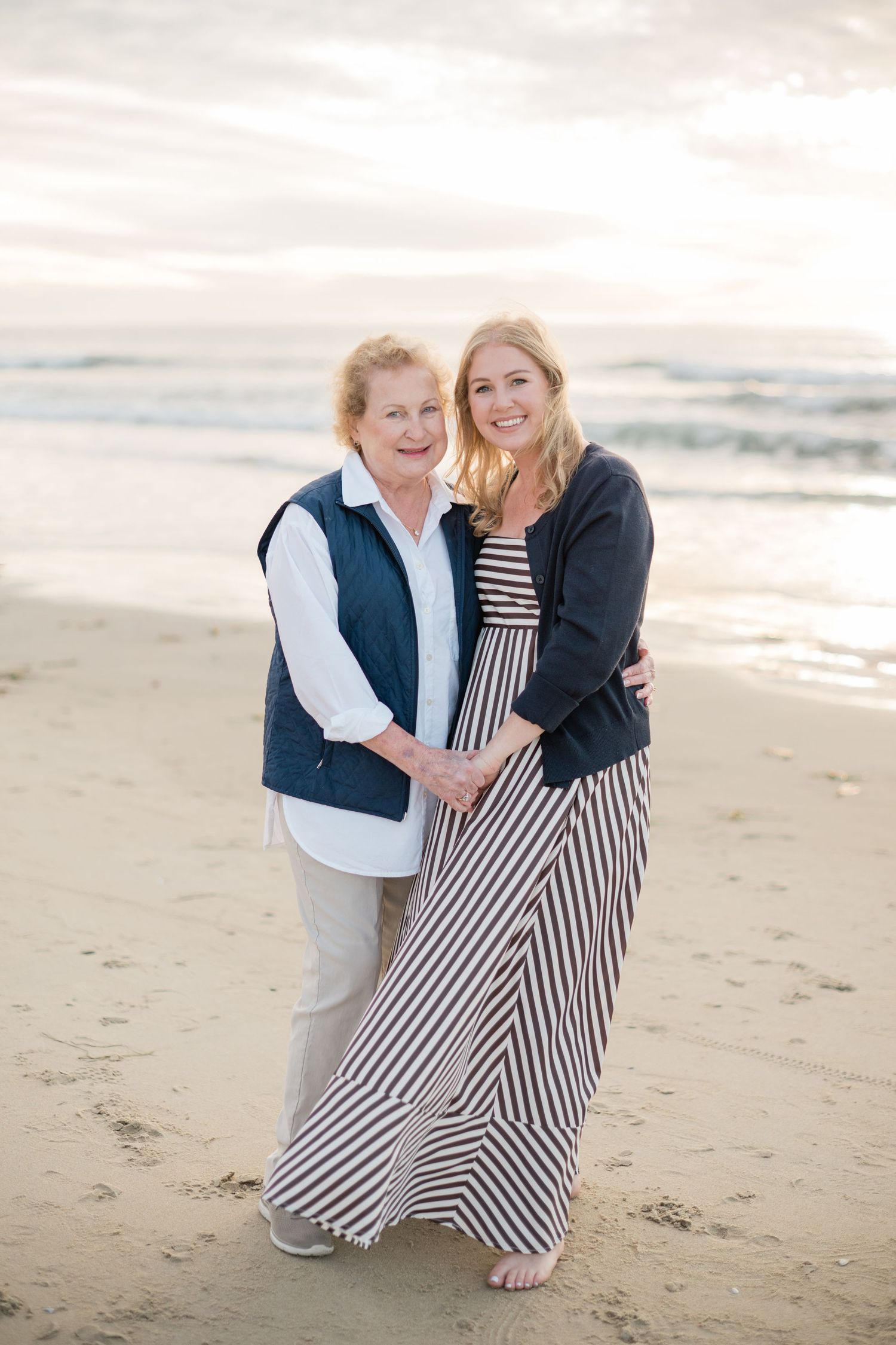 Two women embracing and smiling on a sandy beach at sunset, one older in a blue vest and one younger in a striped maxi dress.