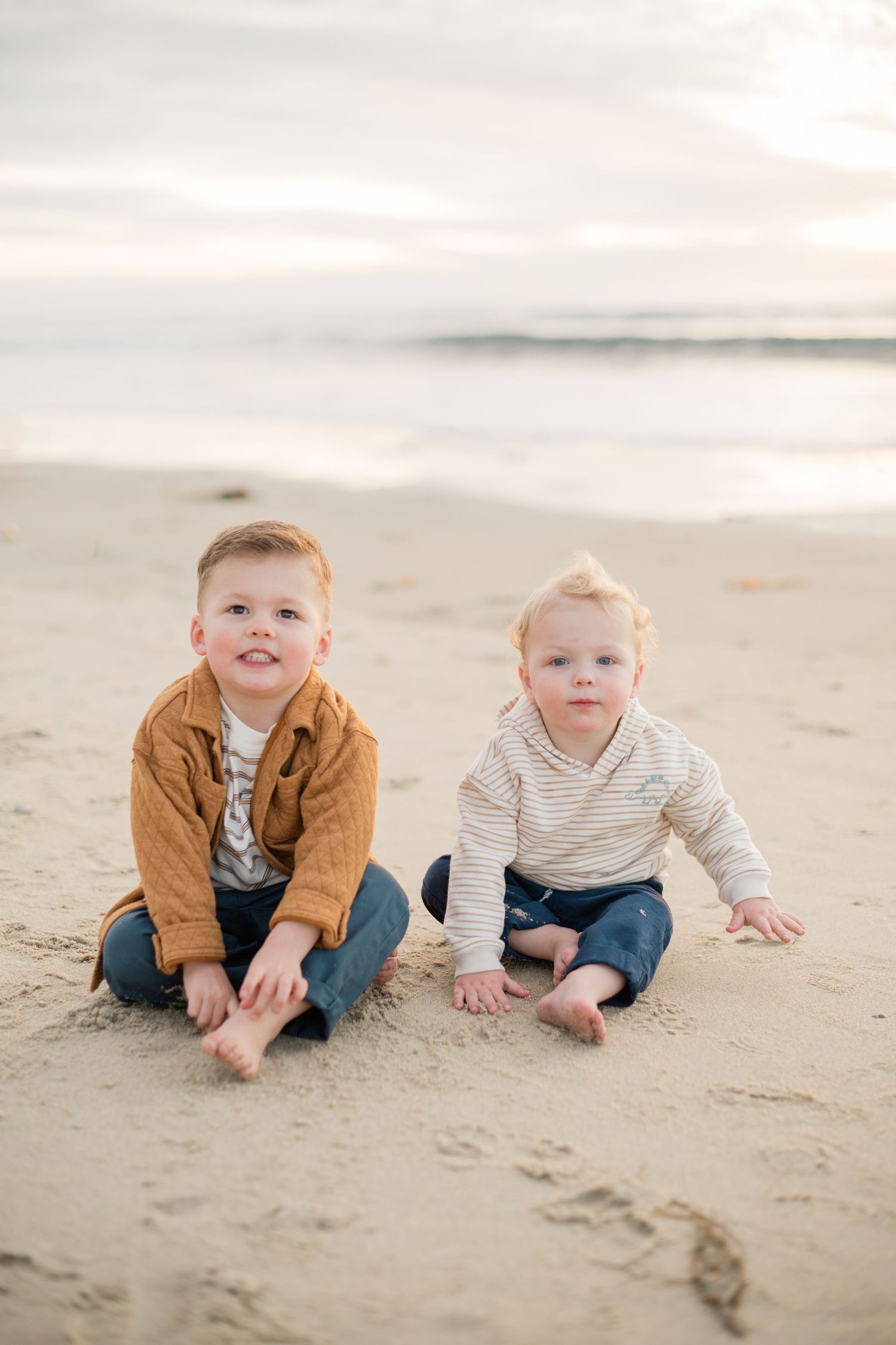 Two young boys sitting on a sandy beach at sunset, wearing casual outfits.