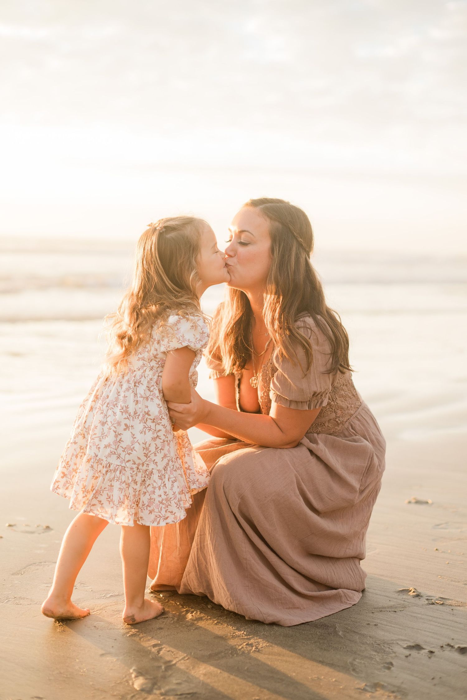 A mother and young daughter share a sweet kiss on the beach at golden hour, both wearing floral dresses.