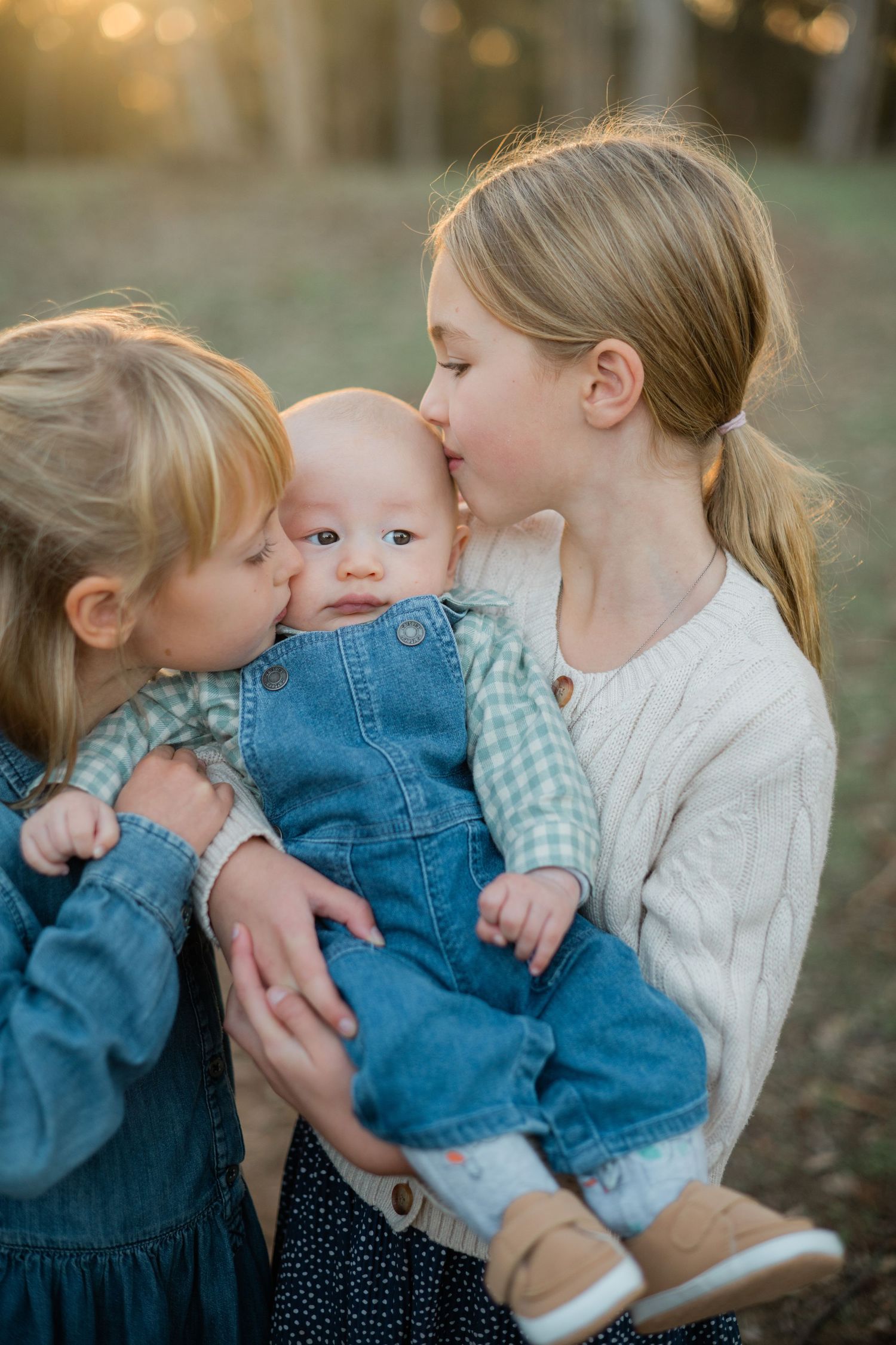 Two blonde sisters lovingly kiss their baby sibling outdoors at golden hour in a heartwarming family portrait.
