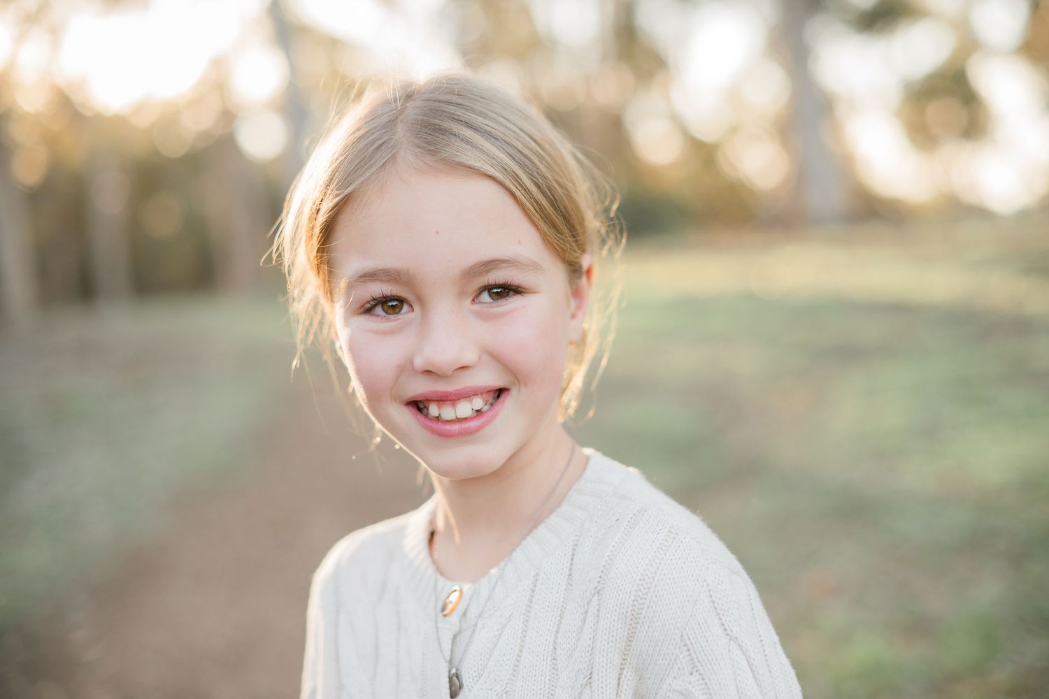 Smiling young blonde girl in a white dress posing outdoors in a sunlit park setting.