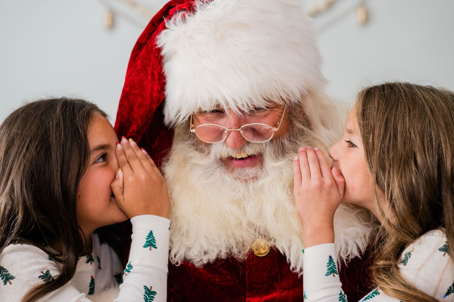 Two girls in Christmas pajamas whispering their holiday wishes to Santa Claus.