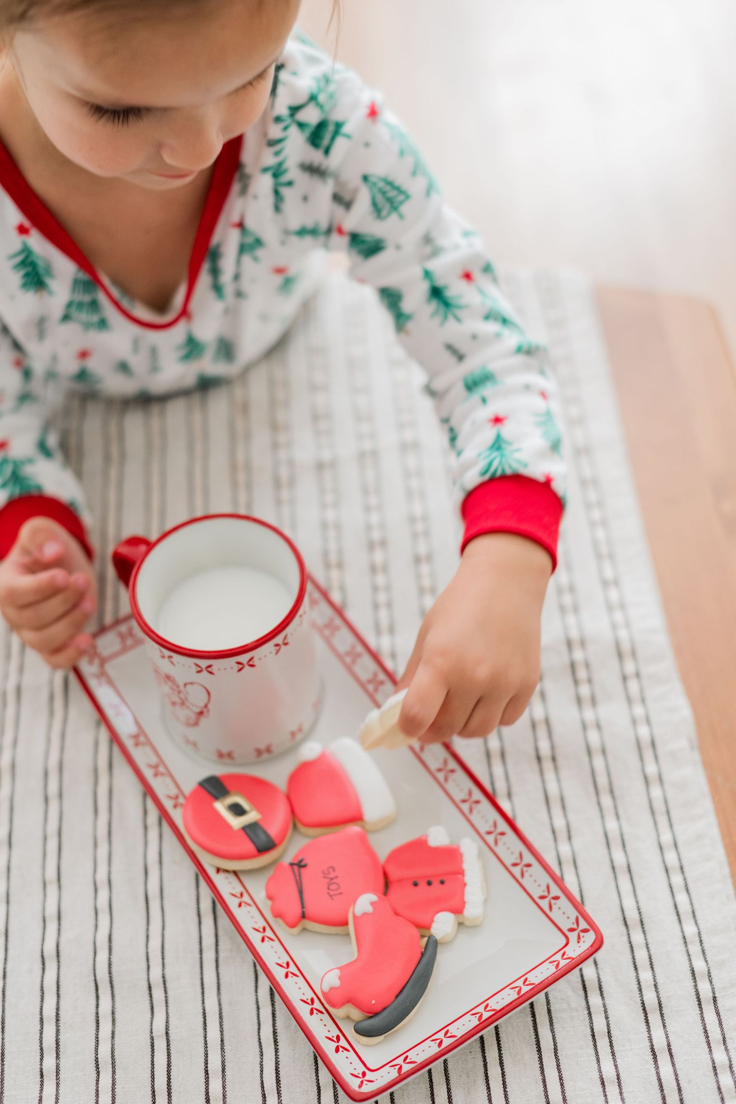 Young girl in Christmas pajamas reaching for Santa-shaped sugar cookies on a holiday tray with a mug of milk.
