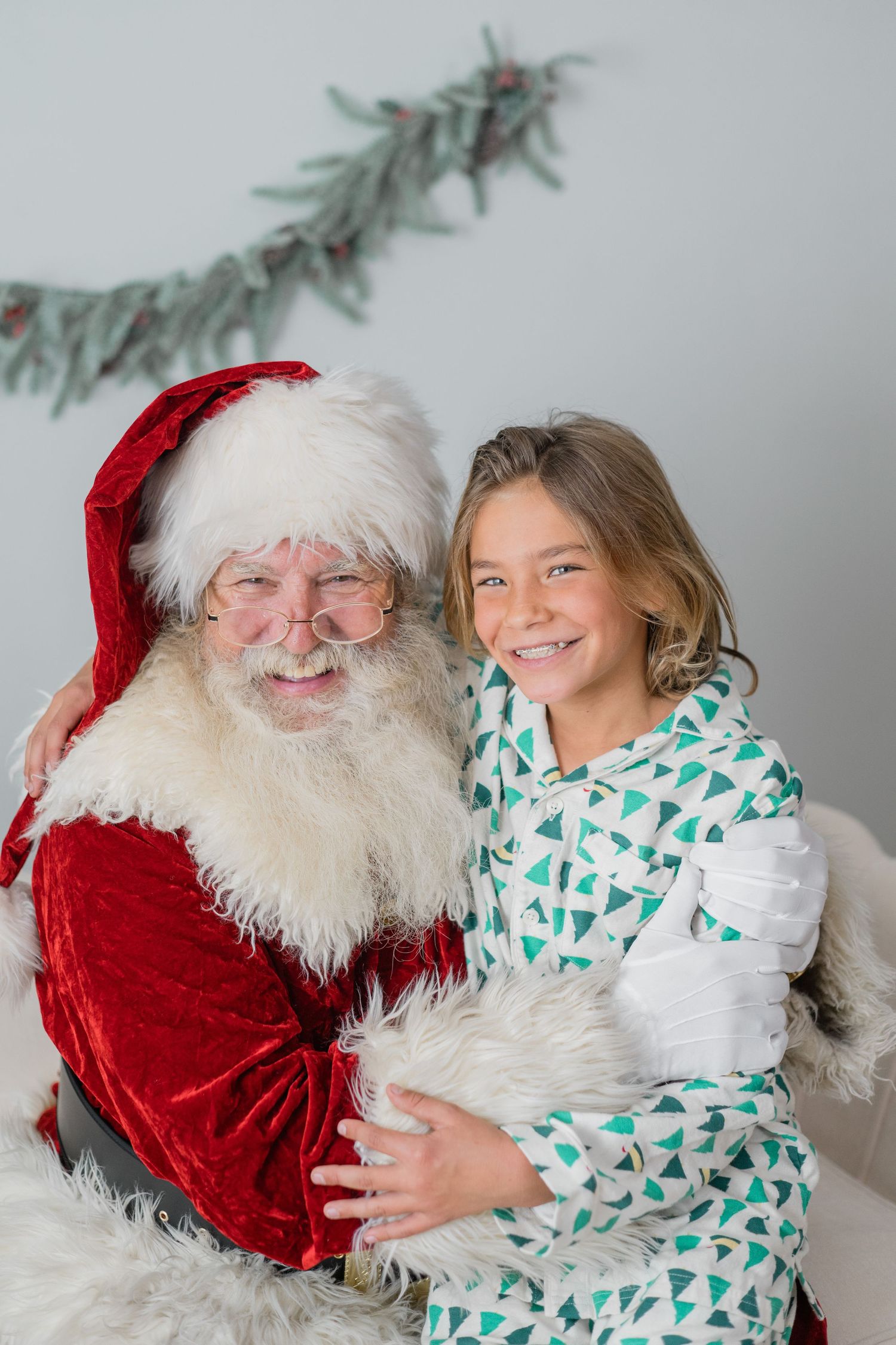 A smiling girl in green pajamas sitting on Santa Claus lap in front of a Christmas garland backdrop.