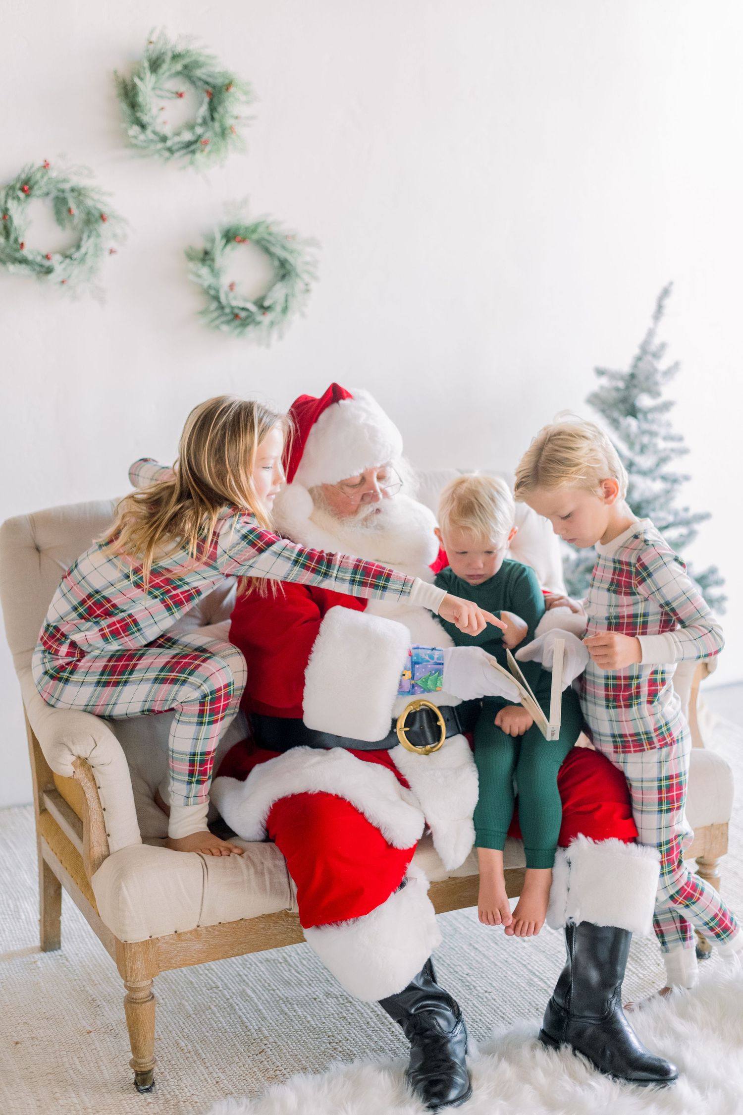 Santa Claus sits on a chair with three children in matching plaid pajamas, reading a Christmas wish list near a decorated tree.