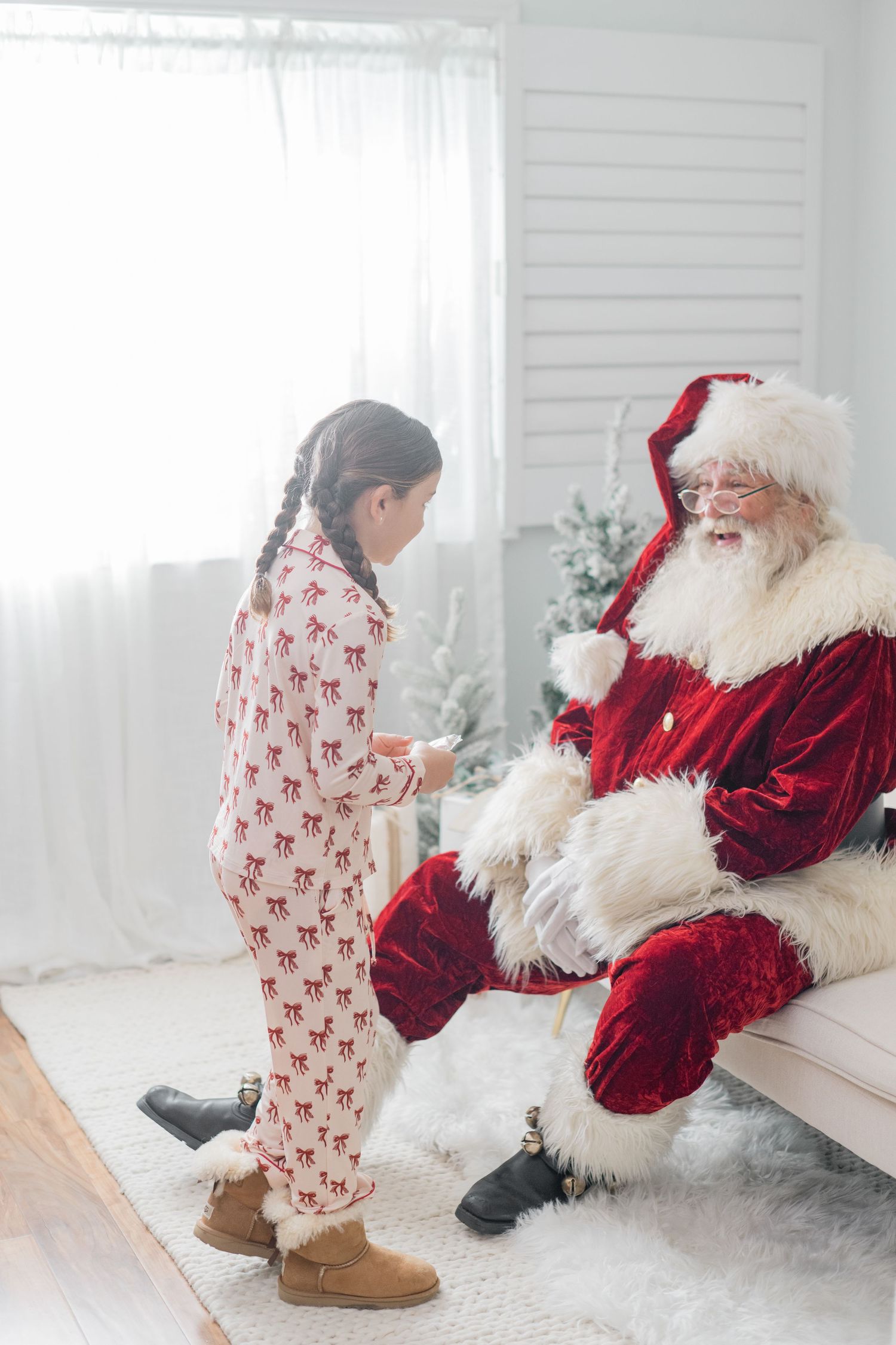 Young girl in reindeer pajamas stands talking to Santa Claus seated in a bright Christmas decorated room.