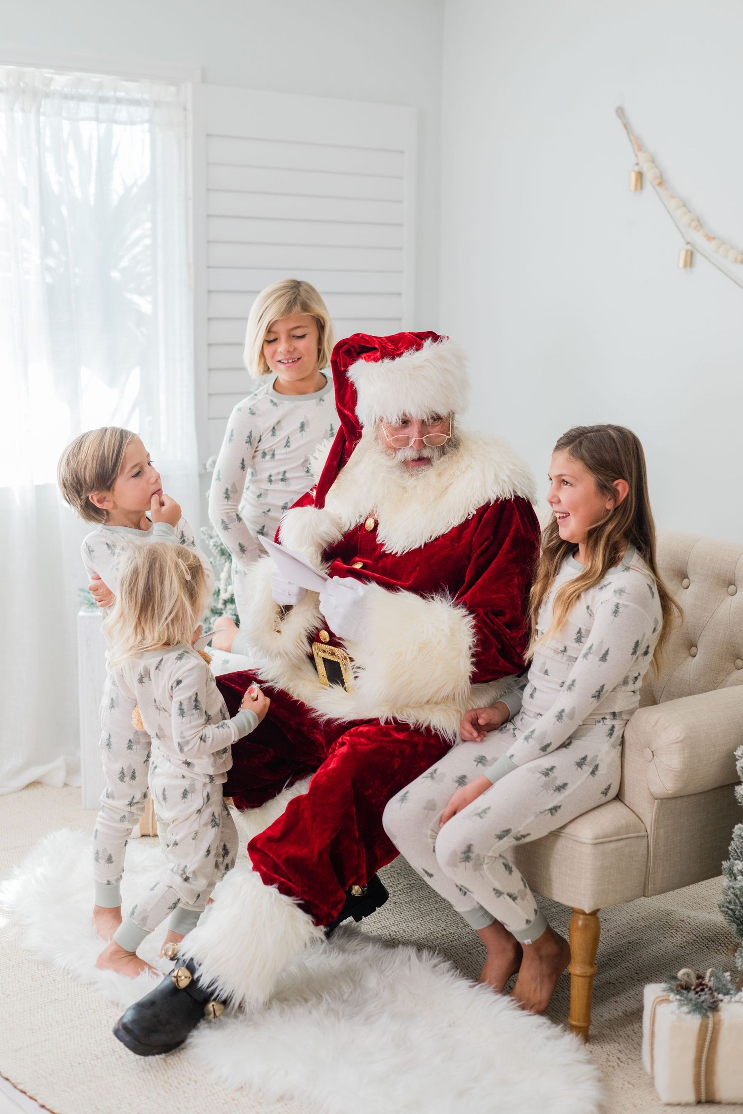 Santa Claus reads his list with three children in matching Christmas pajamas gathered around him in a bright white room.