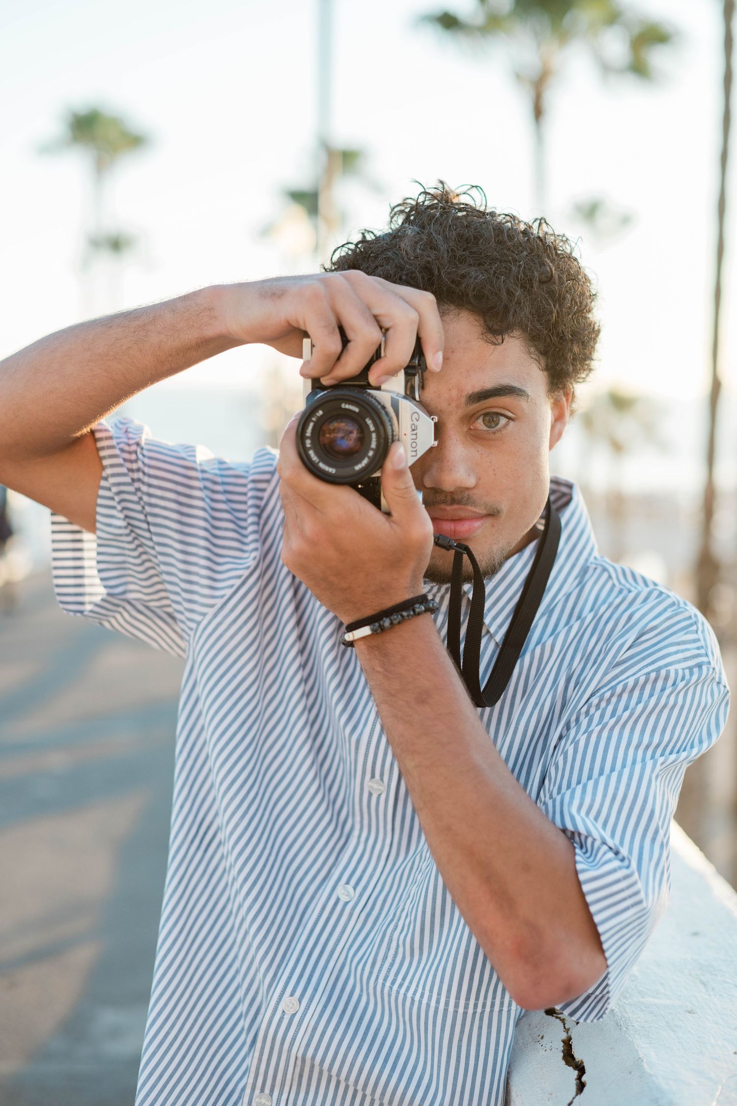 Young man with curly hair holding a film camera outdoors near palm trees, wearing a striped shirt.