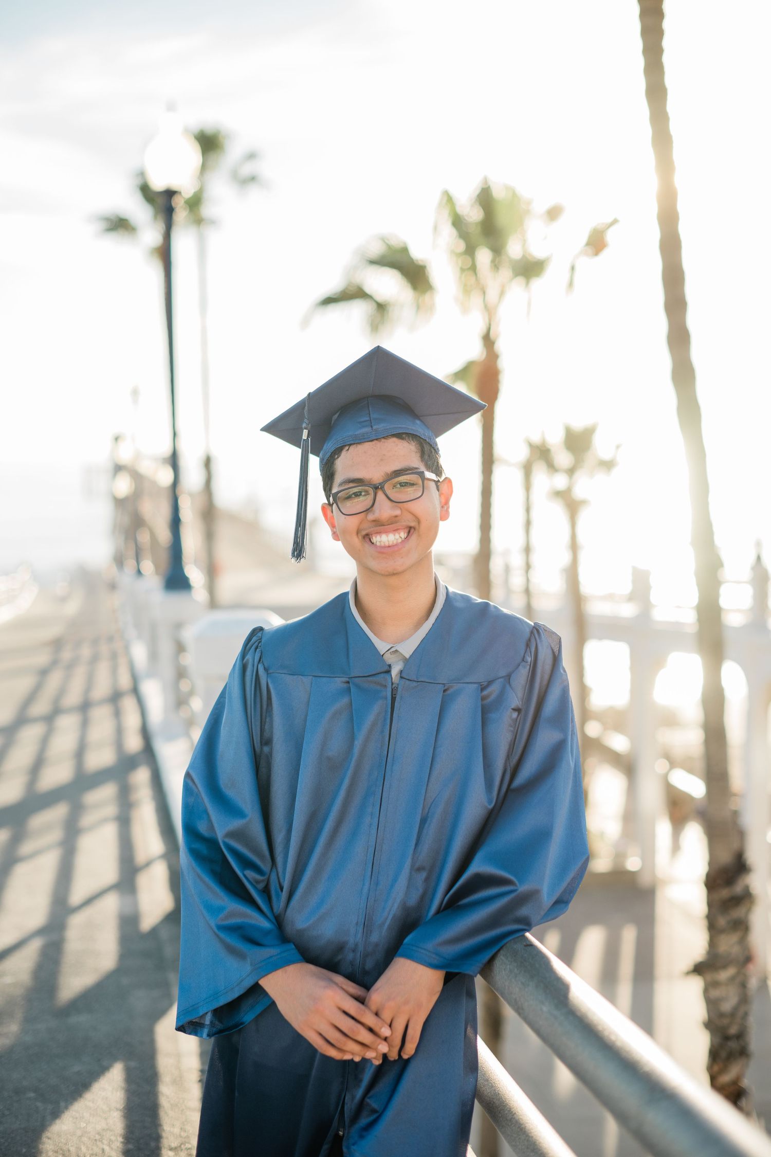 Smiling graduate in blue cap and gown poses on sunny boardwalk with palm trees in background.