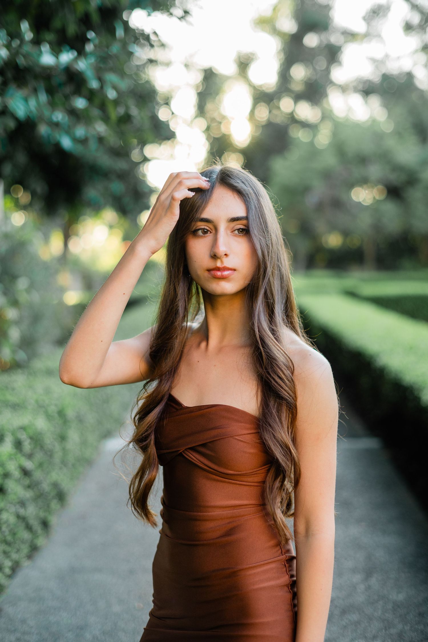 Young woman in brown satin dress poses gracefully in a lush green garden, hand touching her long wavy hair.