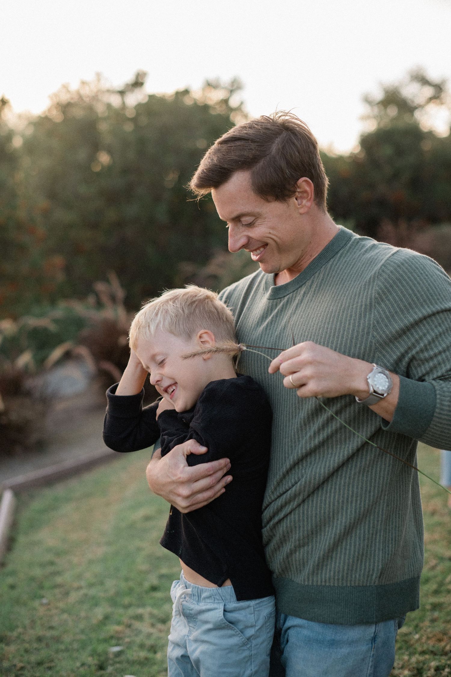 Smiling father hugging laughing young son outdoors in warm golden hour light.