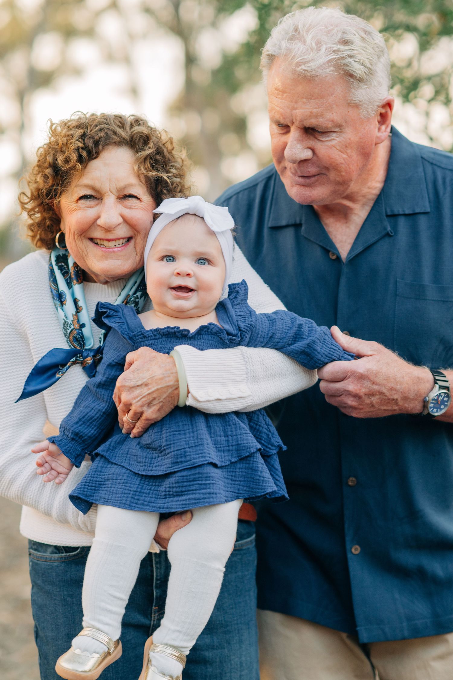Smiling grandparents holding an adorable baby girl in a blue dress outdoors during a family portrait session.
