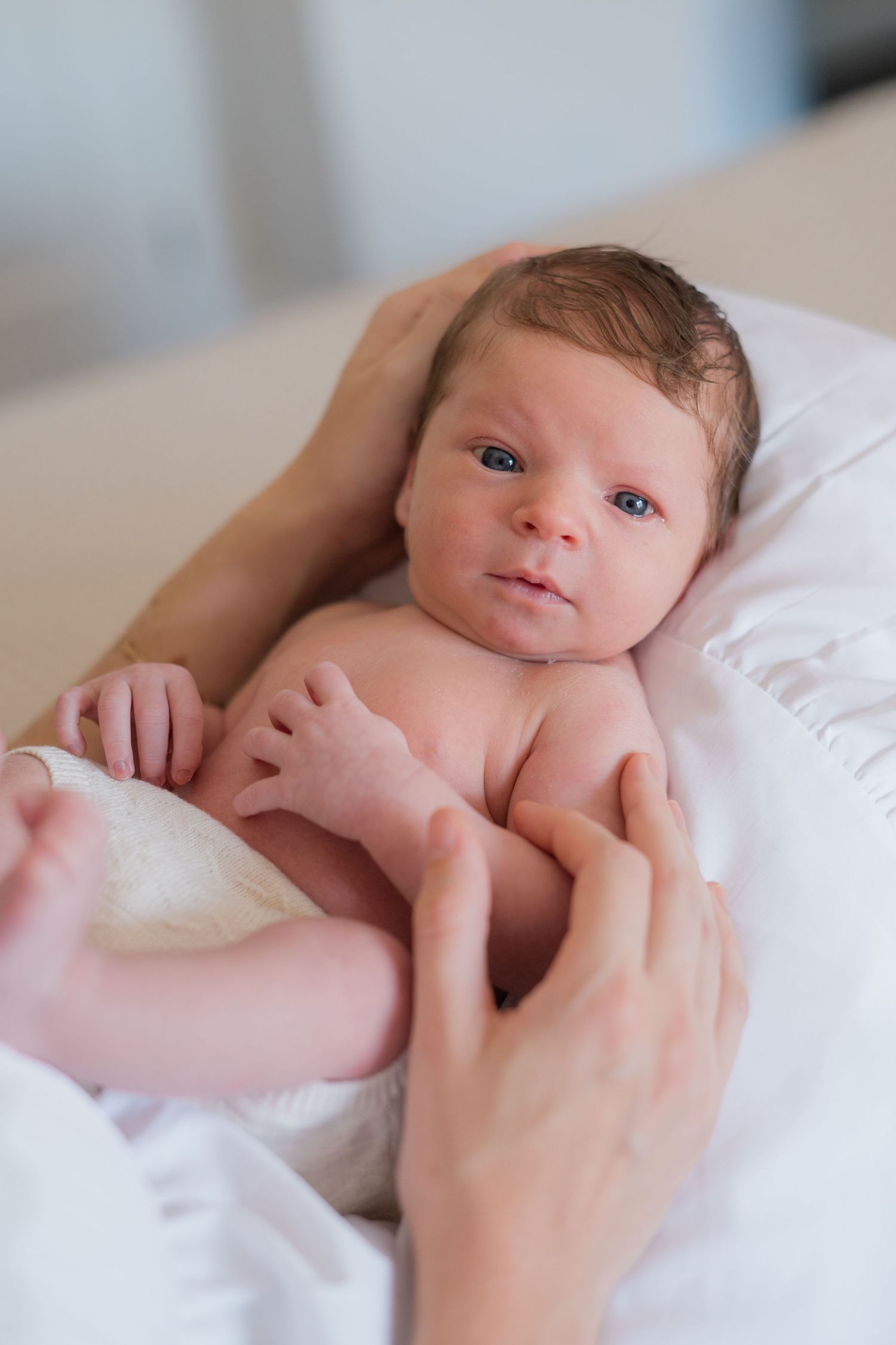 Alert newborn baby with dark hair lying on white blanket held by parent hands.