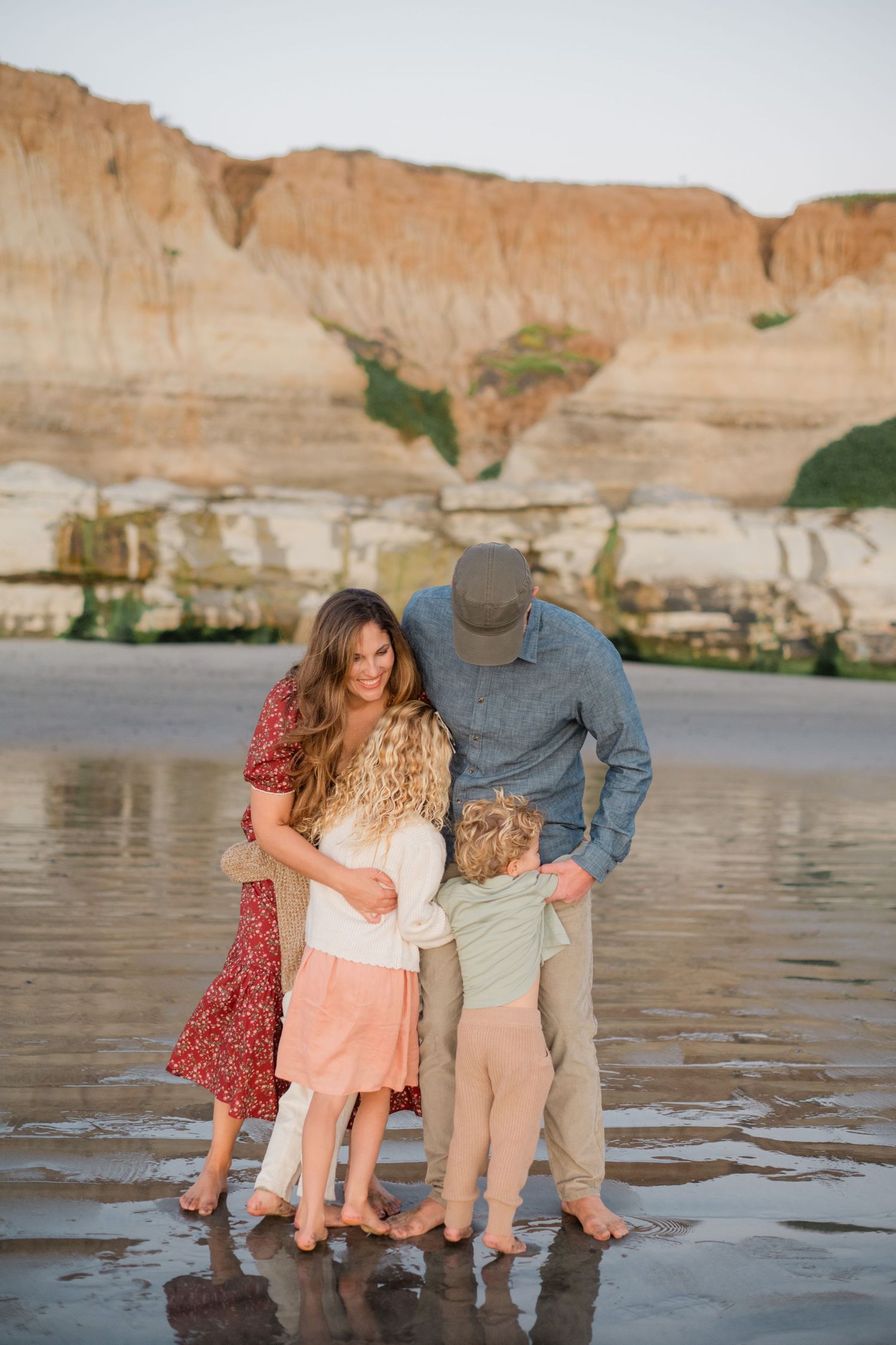 Family of four embracing on a reflective beach with sandy cliffs at golden hour.