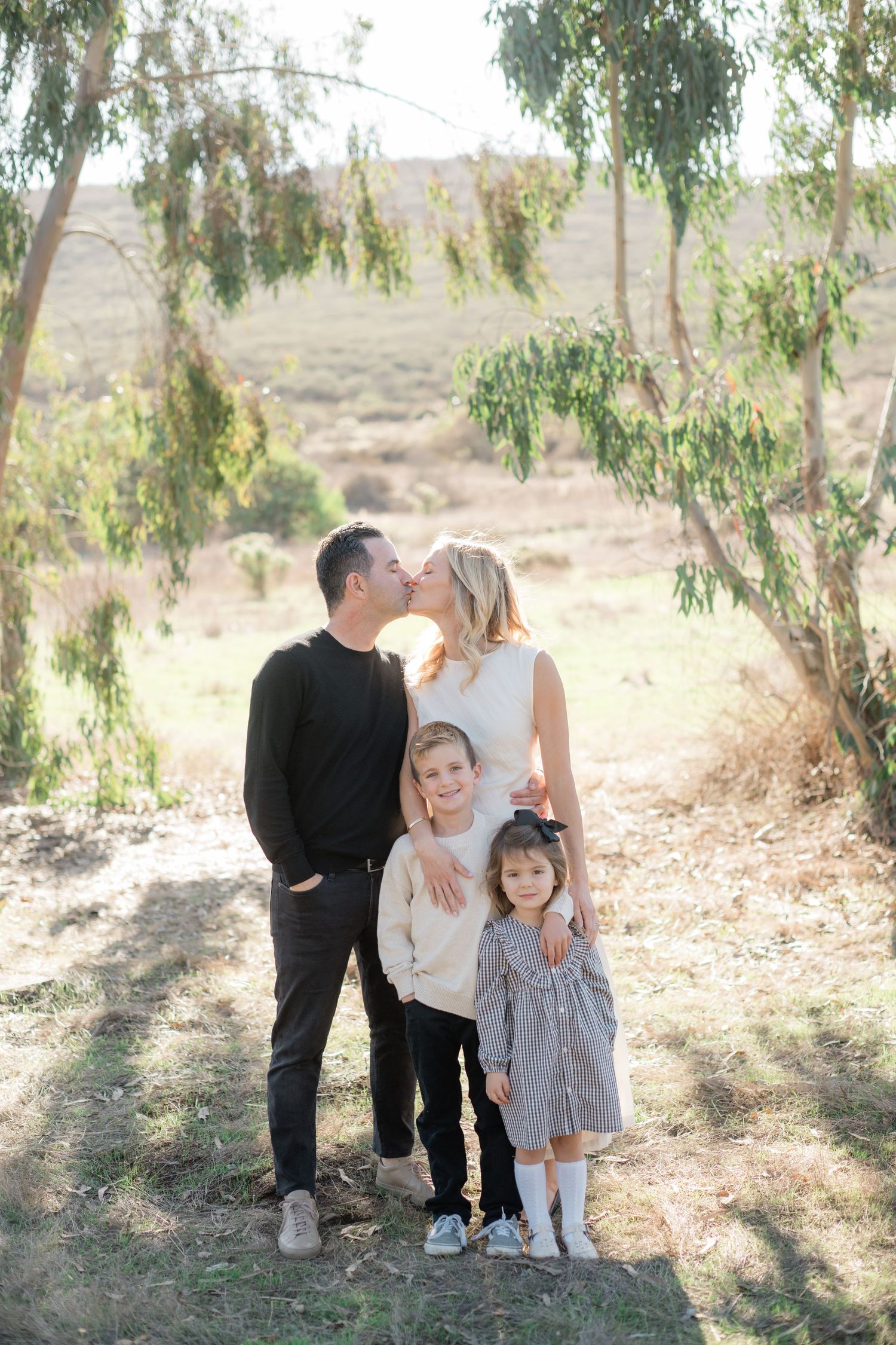 Family of four poses outdoors under eucalyptus trees, parents kissing while children stand in front smiling.