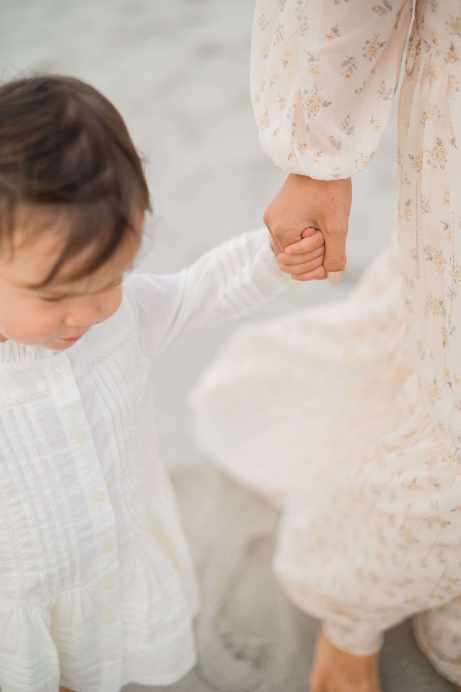 A toddler in a white dress holds hands with a woman in a floral gown on a sandy beach.