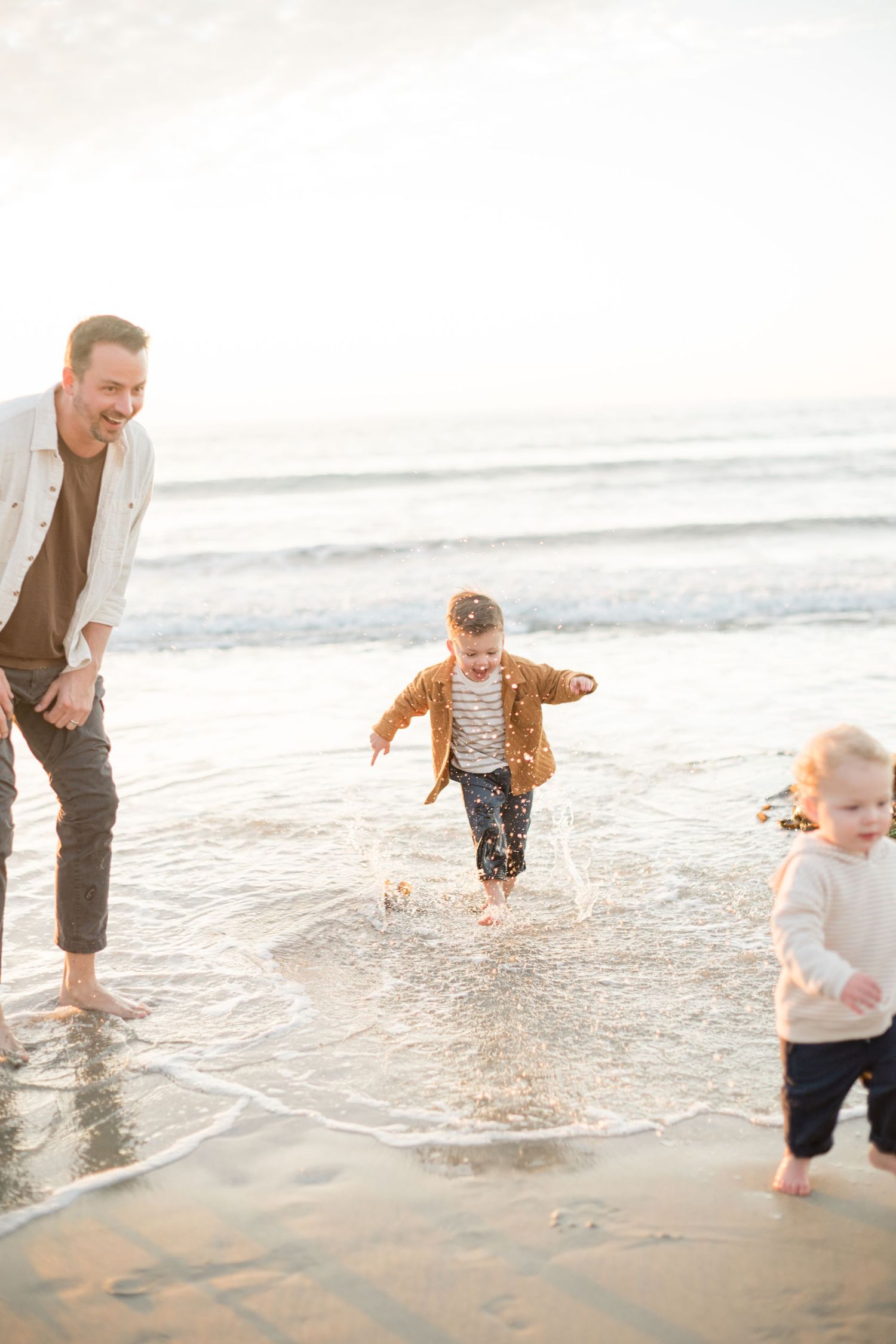 A father watches his young children play in the ocean waves on a sunny beach, splashing in the shallow water.