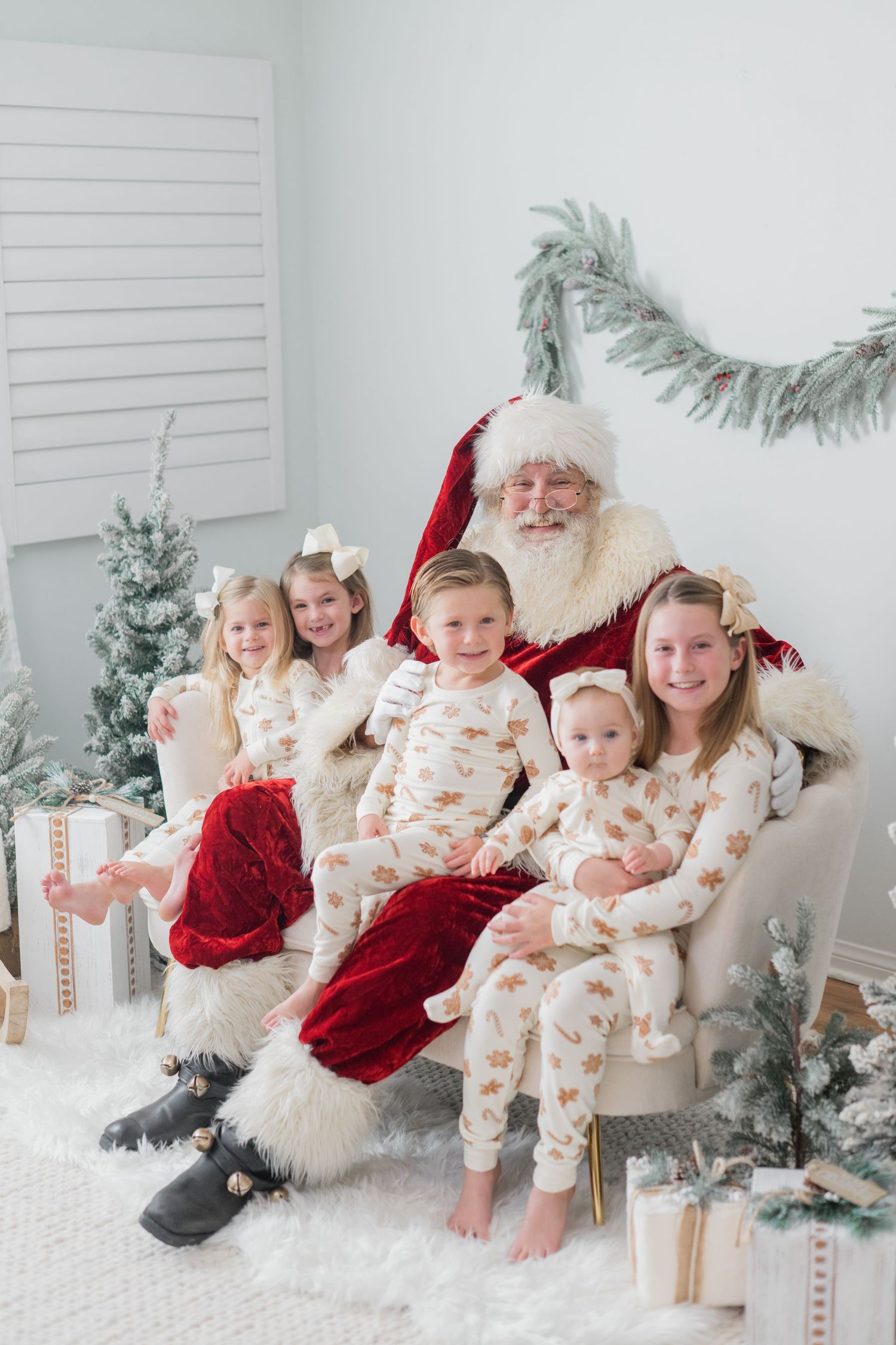 Five children in matching Christmas pajamas pose with Santa Claus on a chair in a festive holiday setting.