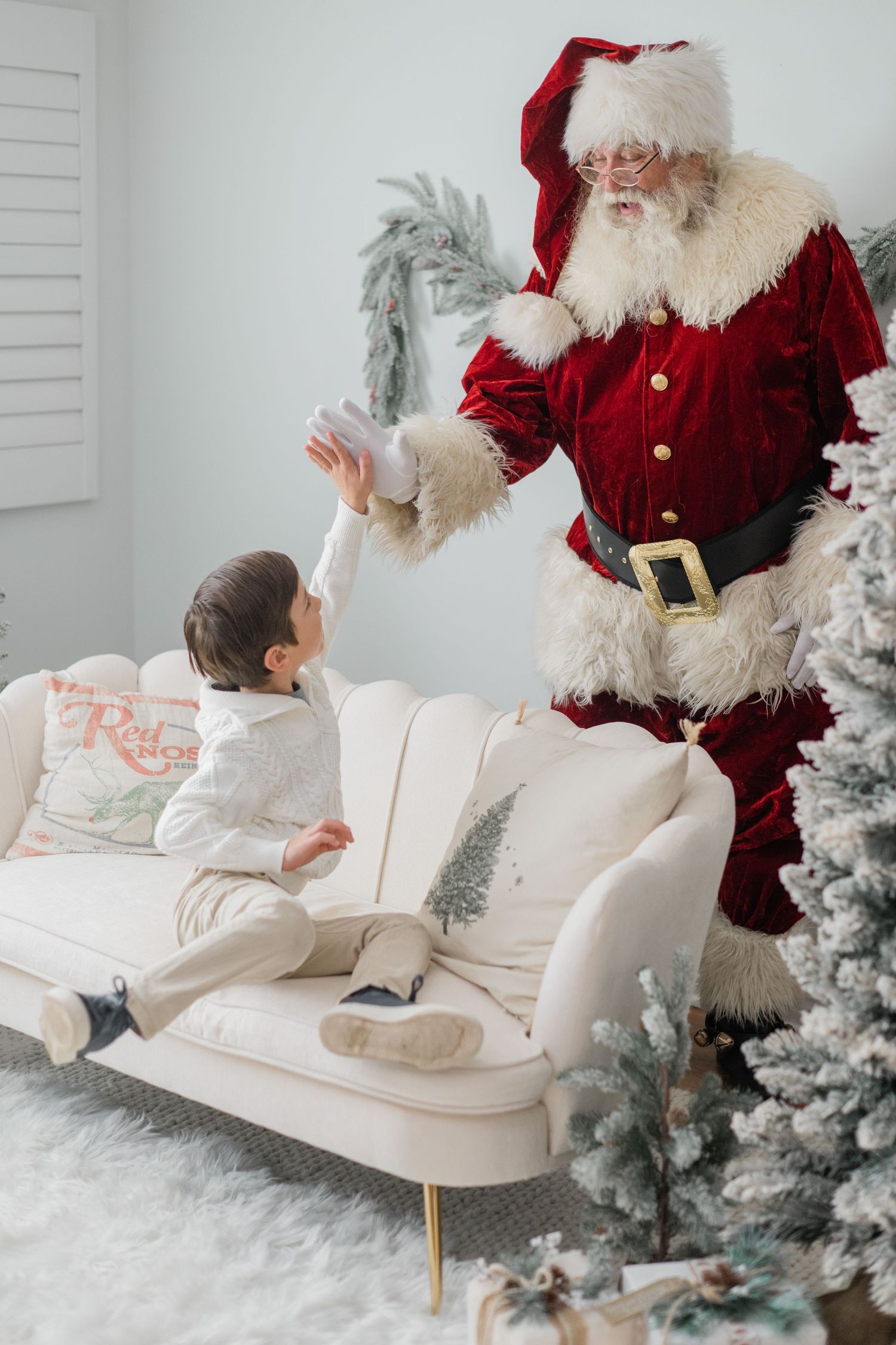 Santa Claus giving a high five to a young child on a white sofa beside a flocked Christmas tree.