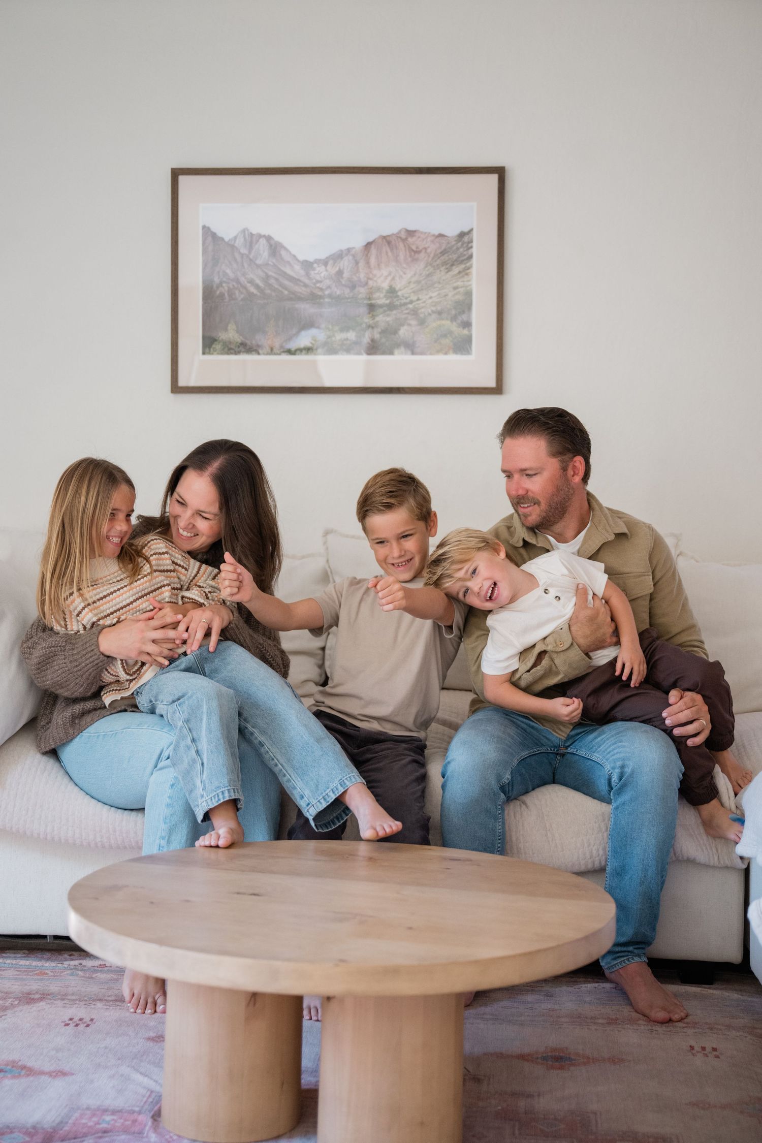 Happy family of five sitting together on a white couch in a bright living room, laughing and playing with a newborn baby.